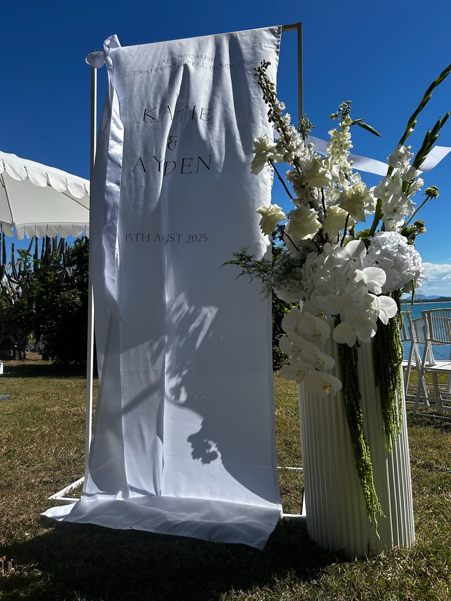 Outdoor wedding setup with a white fabric banner and a floral arrangement of white flowers, including orchids and roses, next to a body of water under a clear blue sky.