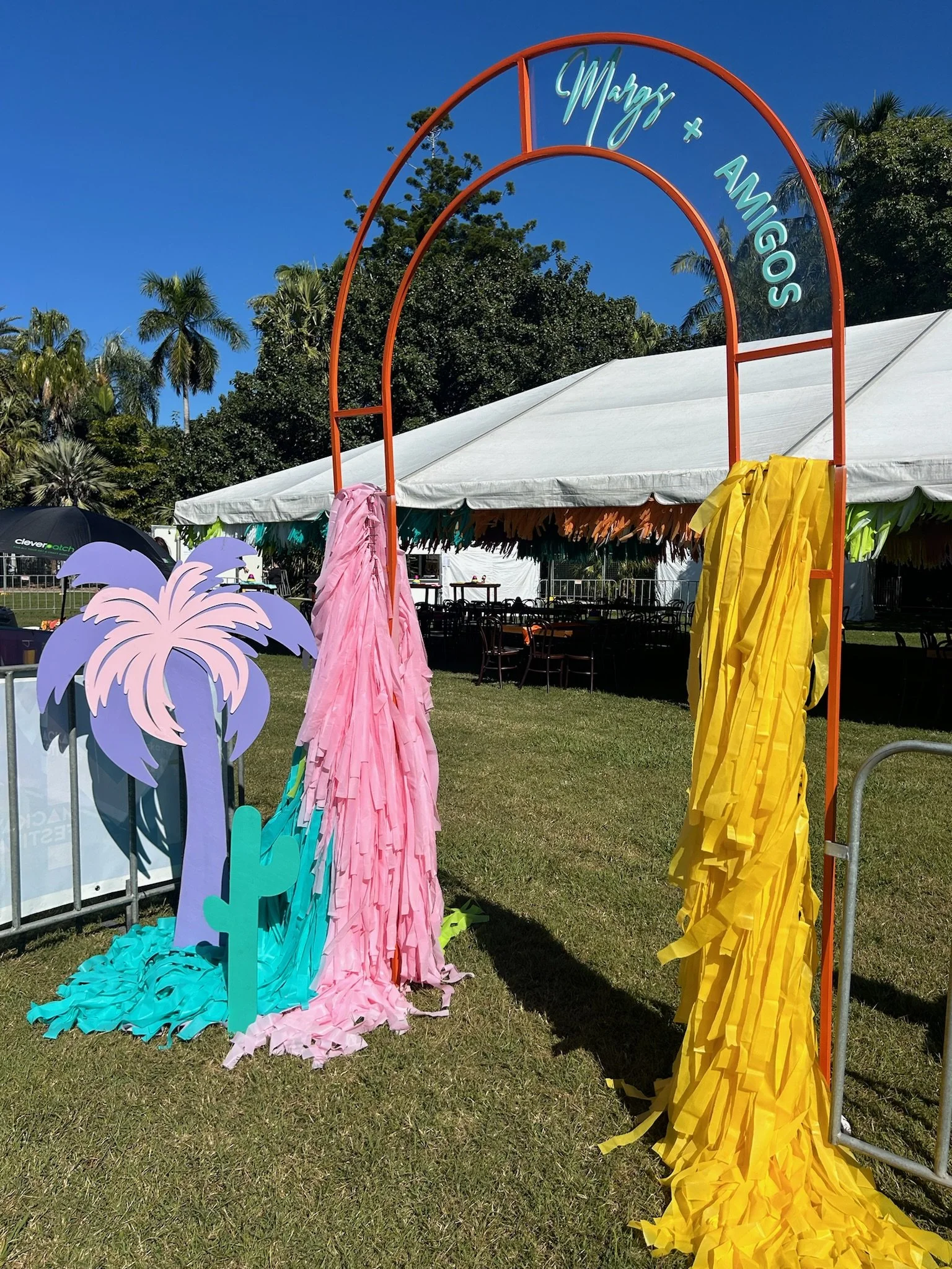 Decorative photo booth arch with signs for 'Marisa + Amigos', featuring hanging pink and yellow fabric strips, a purple palm tree cutout, and cactus decorations, set outdoors under a blue sky with palm trees in the background.