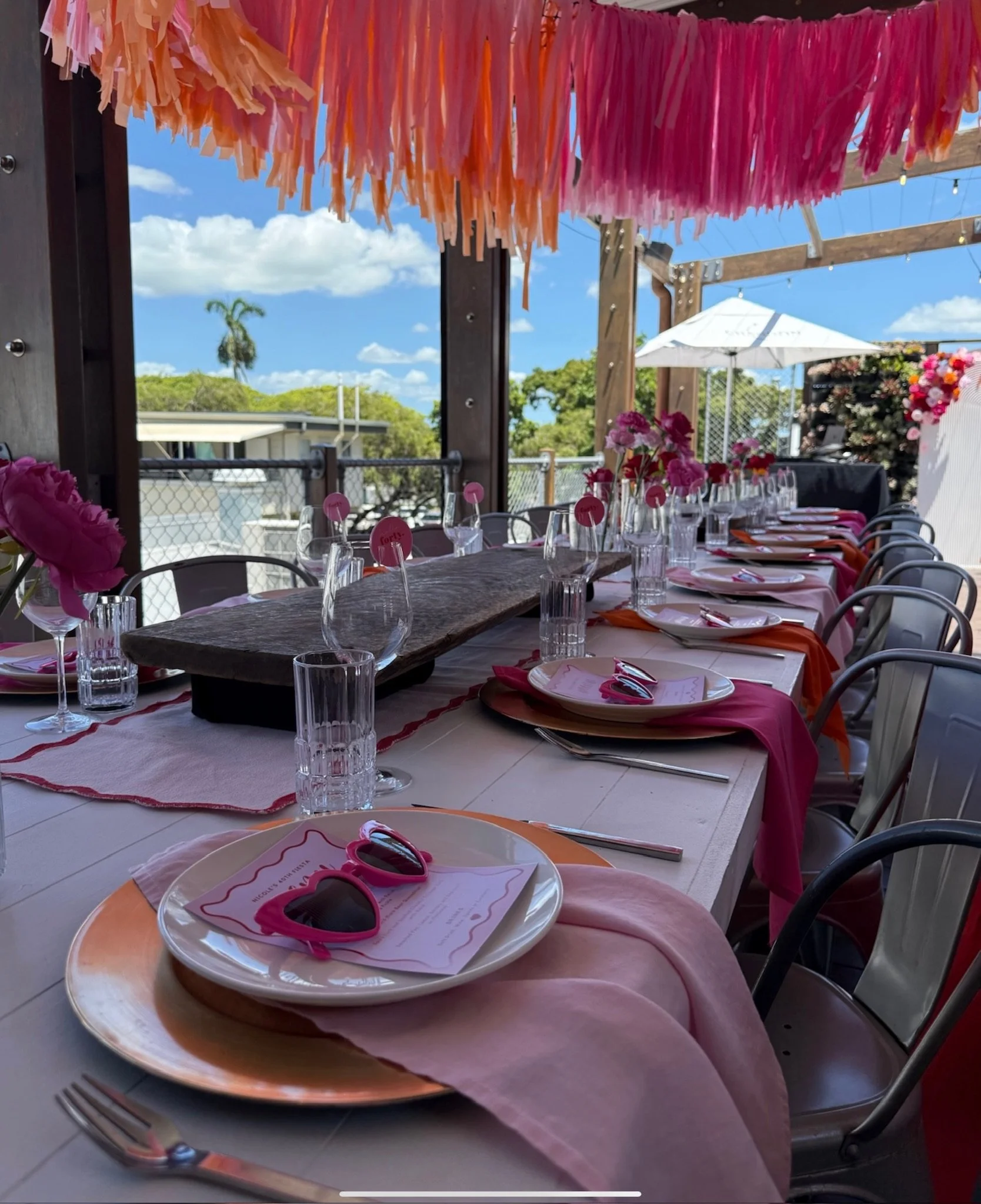 A decorated outdoor dining table setup with pink, orange, and peach accents, pink flowers, pink sunglasses, and a sunny sky with clouds visible in the background.