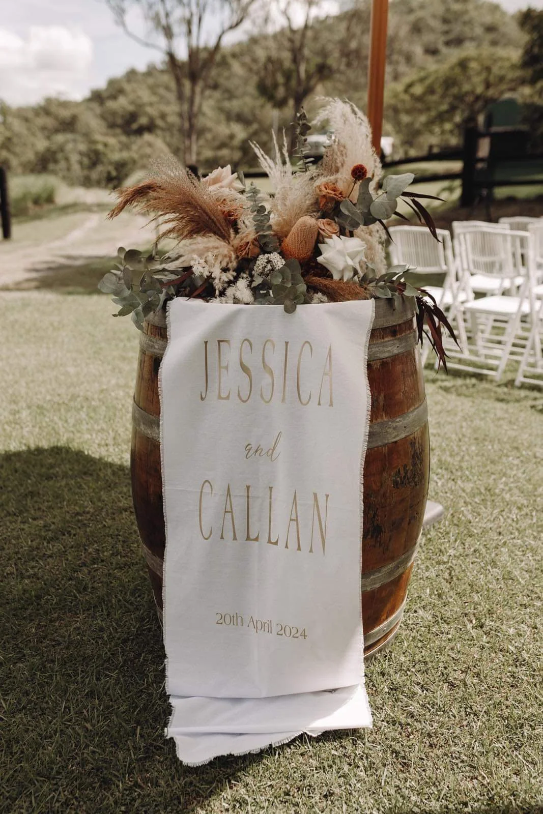 A wooden barrel decorated with flowers and greenery, with a white cloth banner displaying the names Jessica and Callan and the date April 20, 2024, at an outdoor wedding venue.