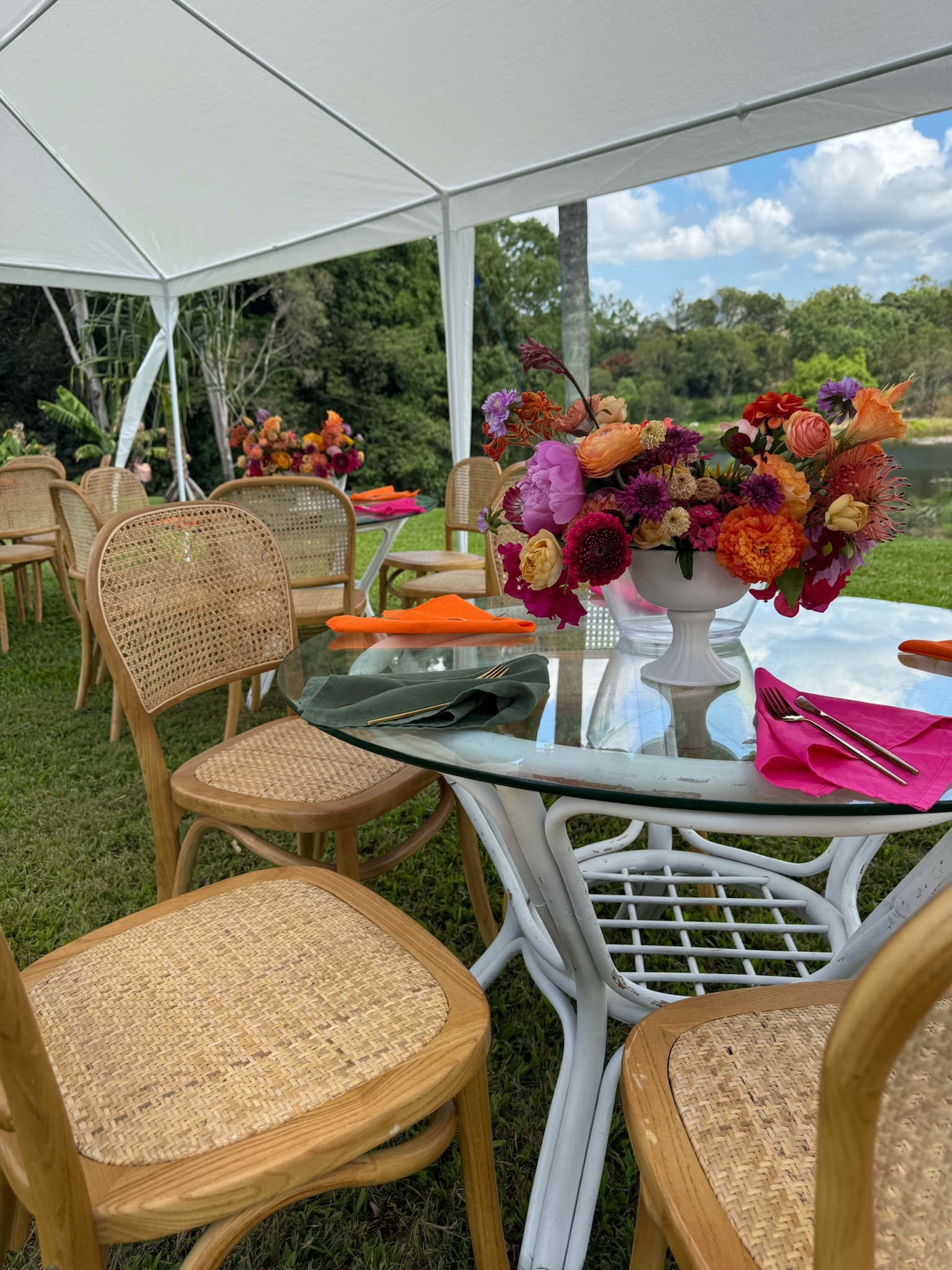 A white outdoor tent set up with a glass table, decorated with a large colorful flower arrangement in a white vase, and hay-colored chairs around the table. The scene is set outdoors with green grass, trees, and a blue sky with clouds in the backgrou