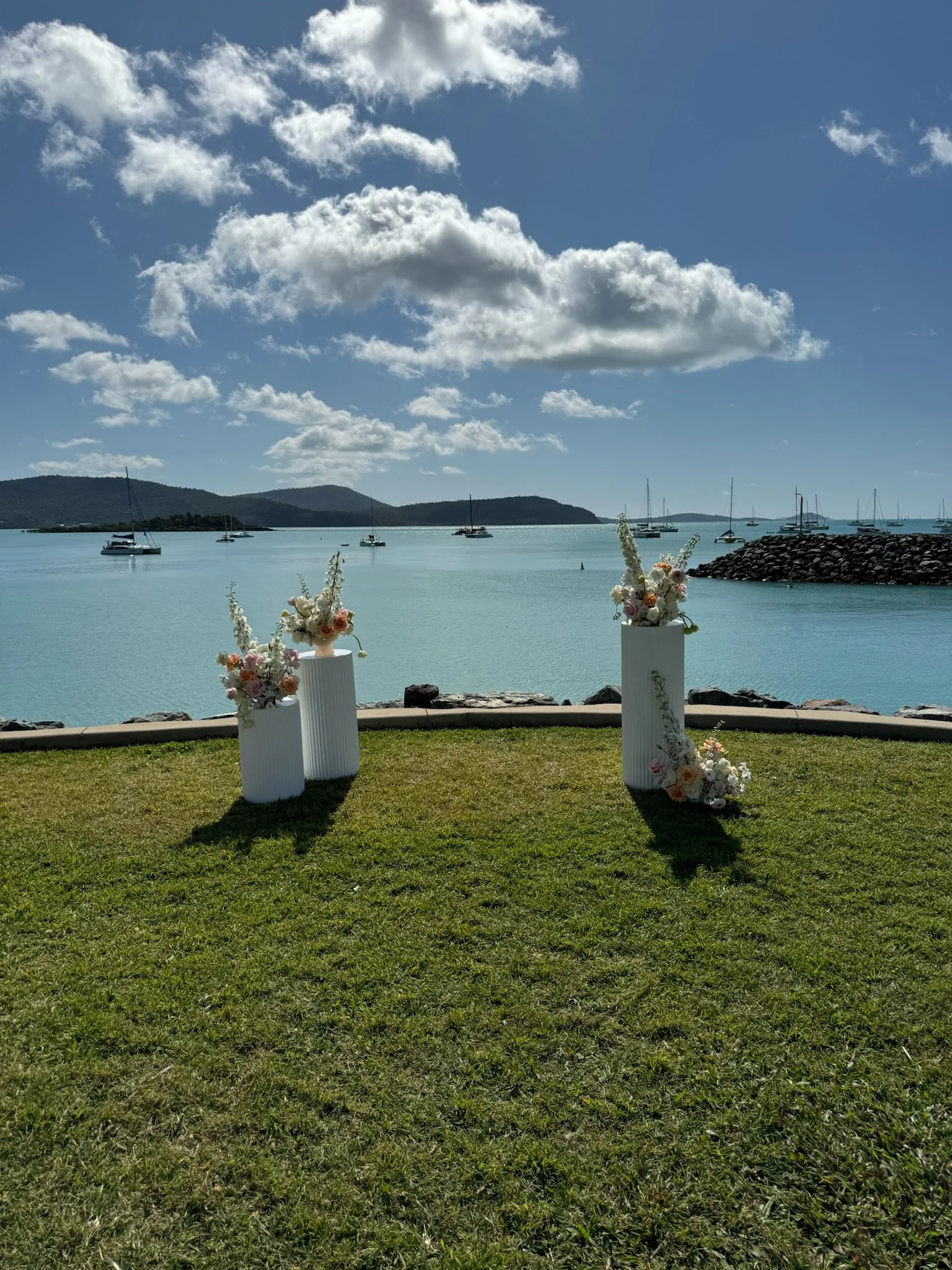 Outdoor wedding setup with three floral arrangements on white pedestals near a body of water, with sailboats and hills in the background and blue sky with clouds.