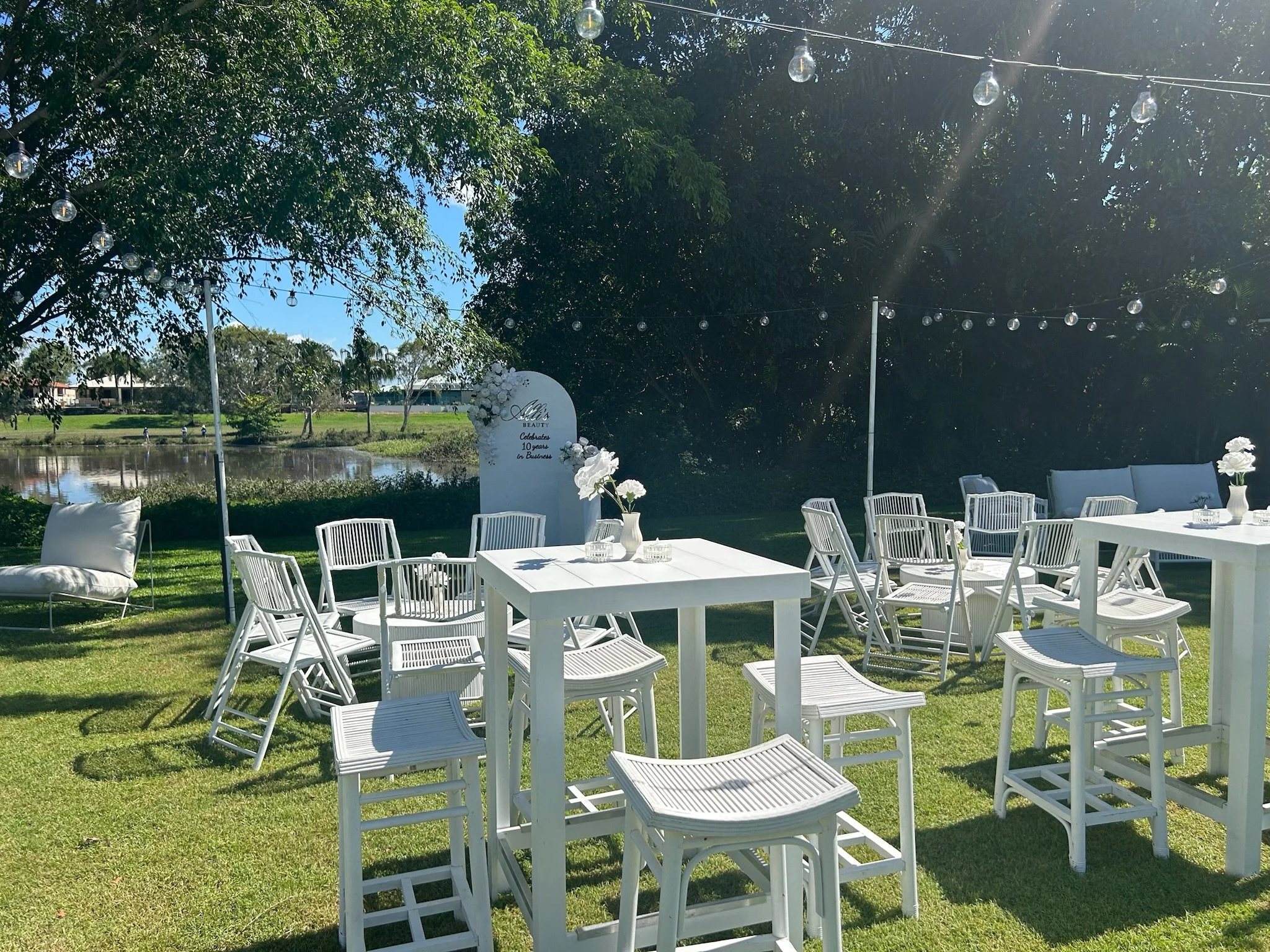 Outdoor event setup with white tables, chairs, and floral centerpieces near a pond, decorated with string lights under a tree on a sunny day.