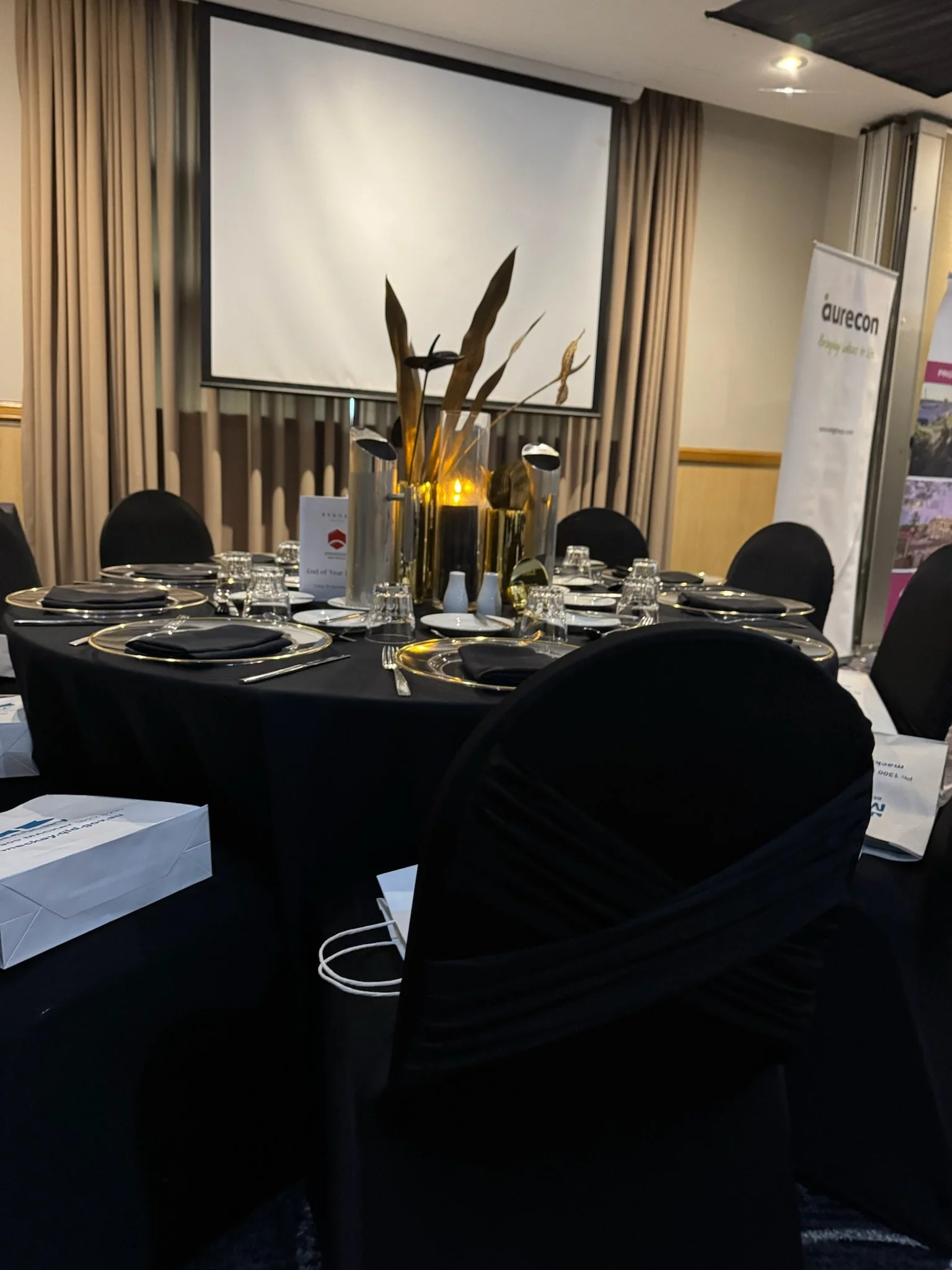 Round banquet table set for a formal event with black tablecloths, black chairs, and gold and silver tableware and decorations. There is a white box on the table and a large gold vase with tall, decorative leaves and black and yellow accents as a cen