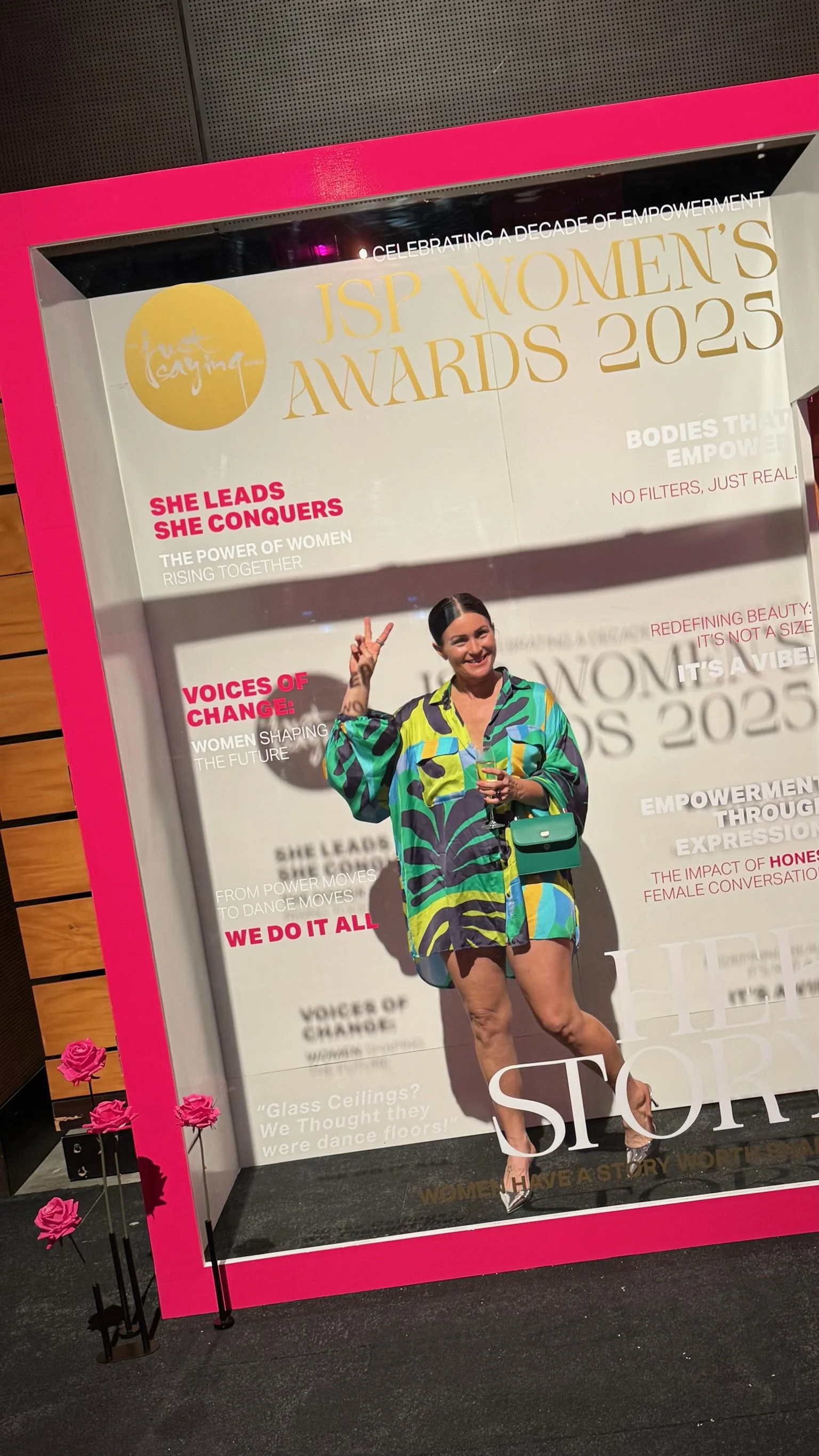 A woman posing inside a decorative photo frame at the JSP Women's Awards 2023 event, holding a drink, wearing a colorful patterned dress, with pink roses on the floor beside the frame.