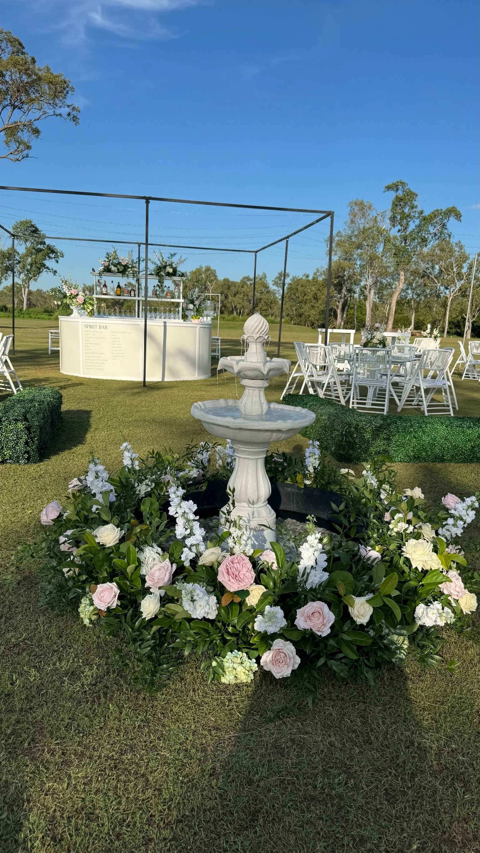 Outdoor wedding venue with a white fountain surrounded by flowers in the foreground, a striped bar under a metal frame, and round tables with white chairs on green grass with trees in the background.