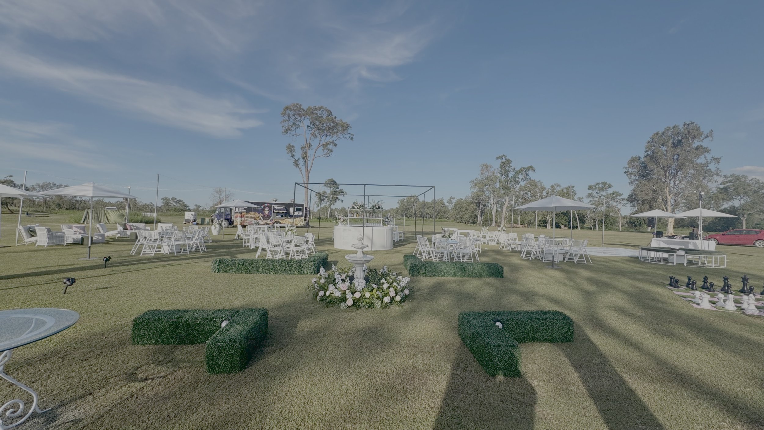 Outdoor event setup on a grassy field with several white tables and chairs, large shade umbrellas, decorative hedges, a chessboard, trees, and a blue sky with clouds.