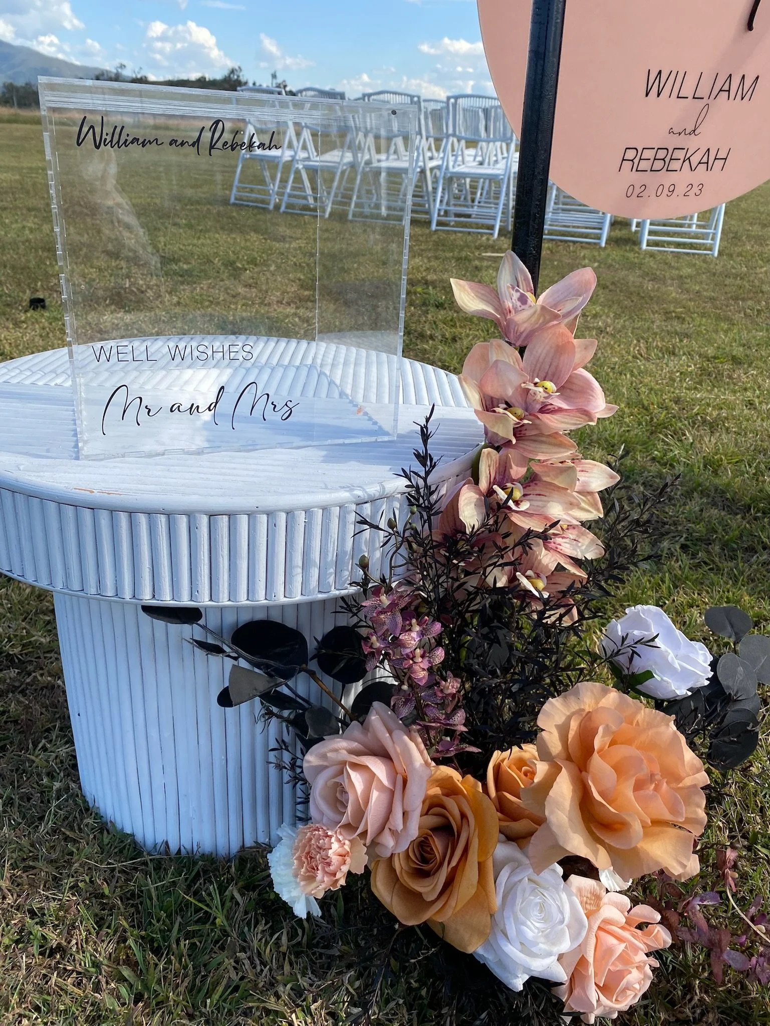 A wedding sign display with a clear acrylic sign that reads 'William and Rebekah, Well Wishes, Mr and Mrs' placed on a white textured table, decorated with pink, peach, white, and purple flowers and greenery, outdoors on grass with stacked white chai