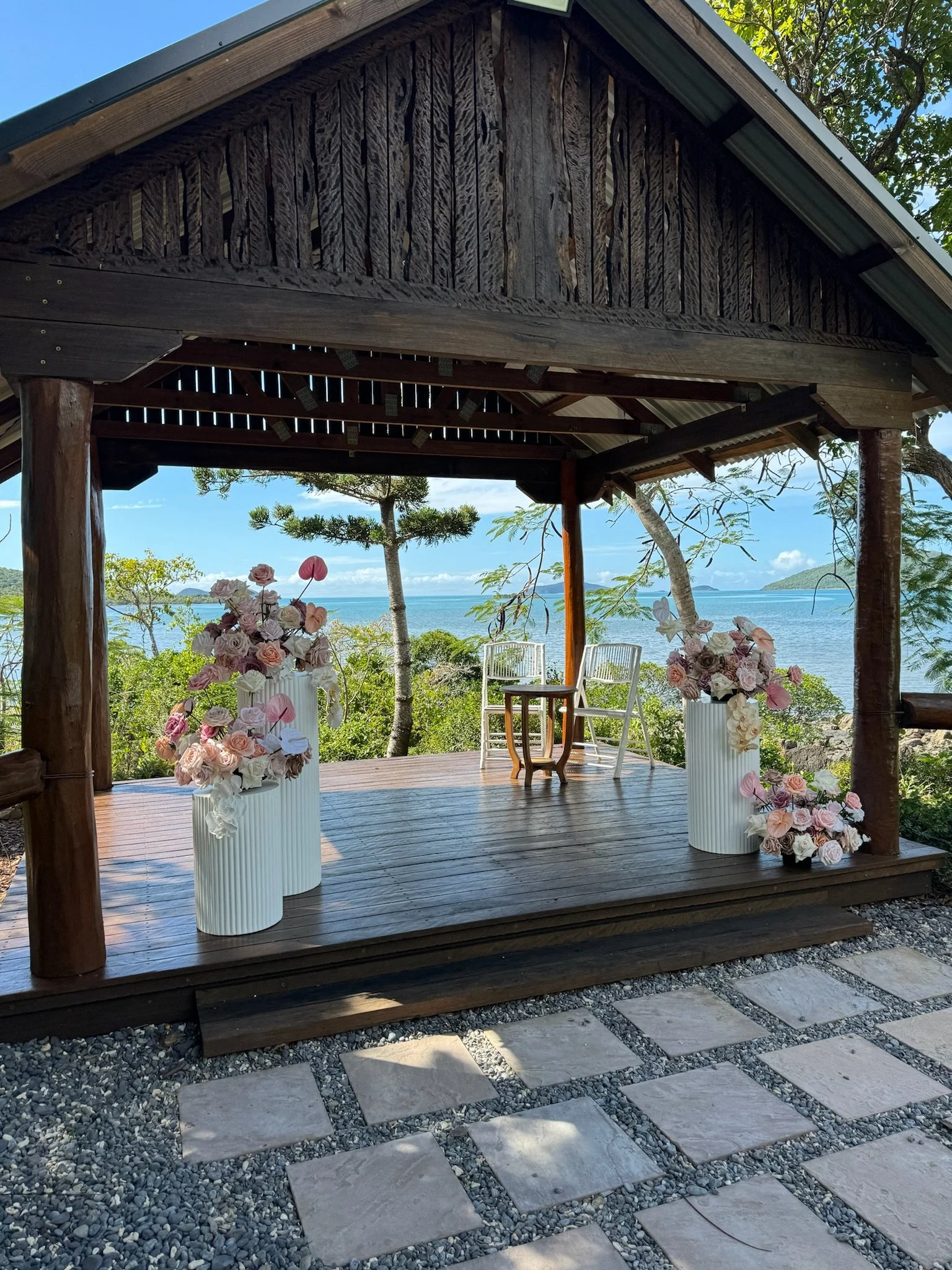 A small outdoor stage decorated with large white vases filled with pink and white flowers, overlooking a scenic ocean view with trees and islands in the background.