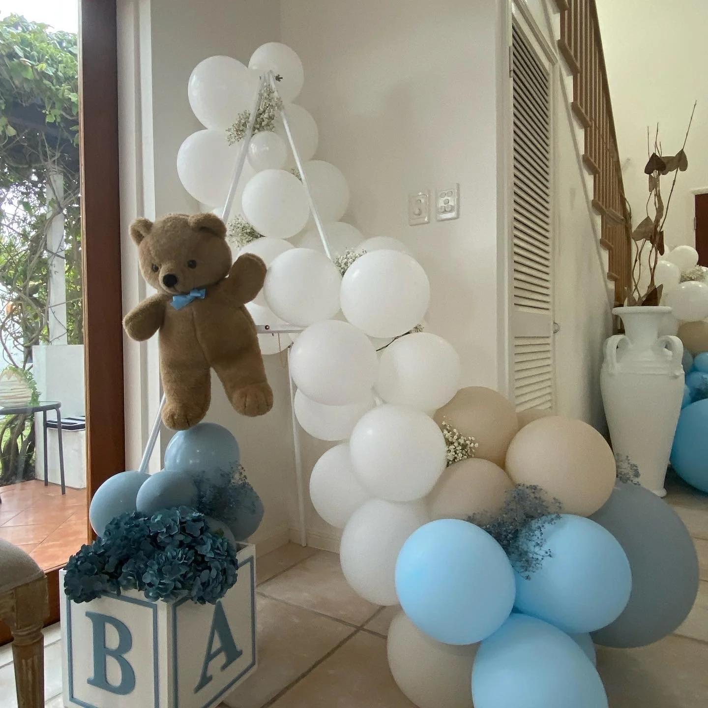 Balloon display with white, cream, and blue balloons, a hanging teddy bear with a blue bow tie, a white box filled with dark blue flowers, and decorative blocks with letters 'B' and 'A' in front of a glass door with greenery outside.