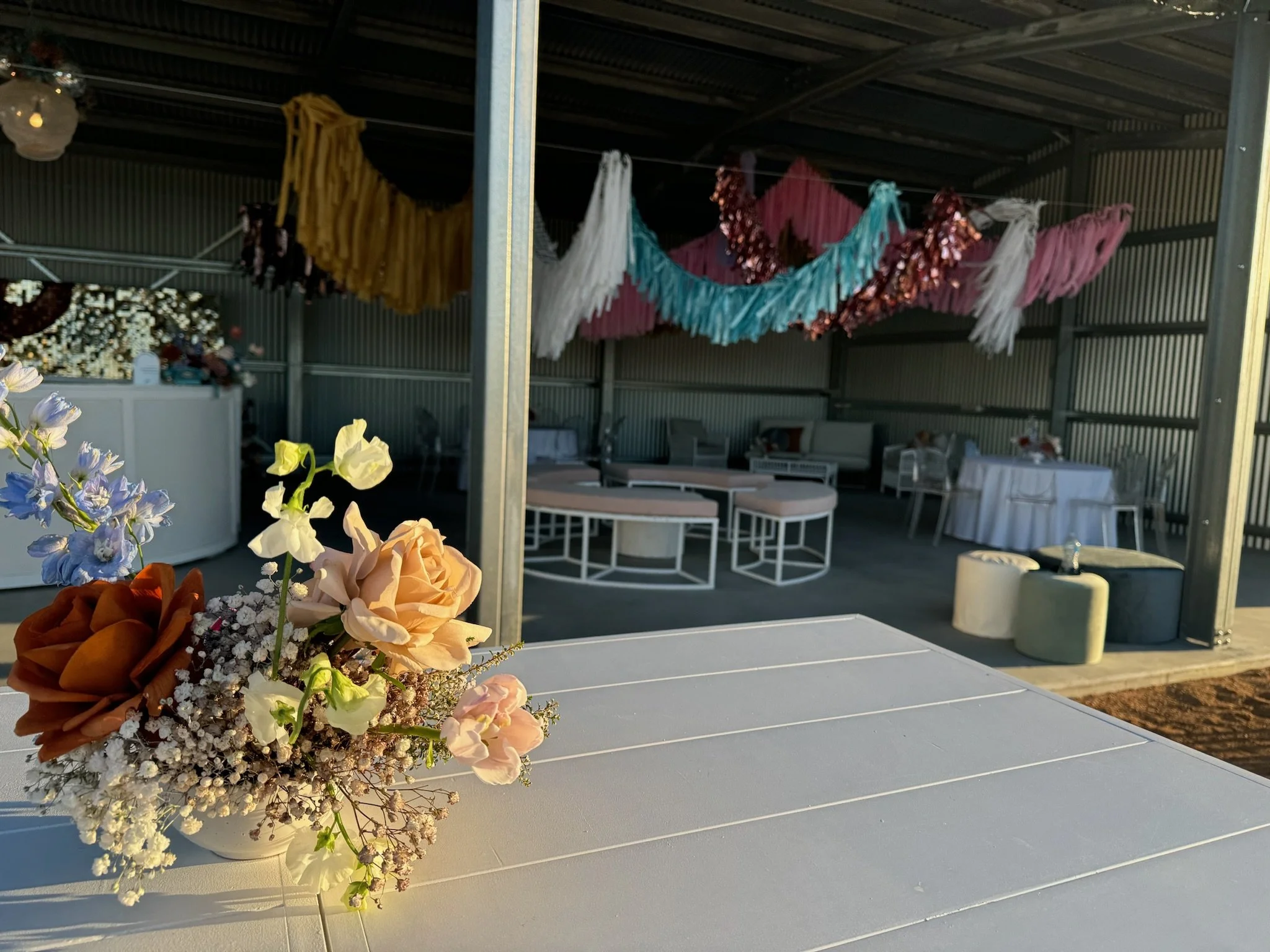 A floral arrangement with roses and other flowers on a white table in the foreground, with a decorated event space featuring colorful hanging fabrics and seating area in the background.