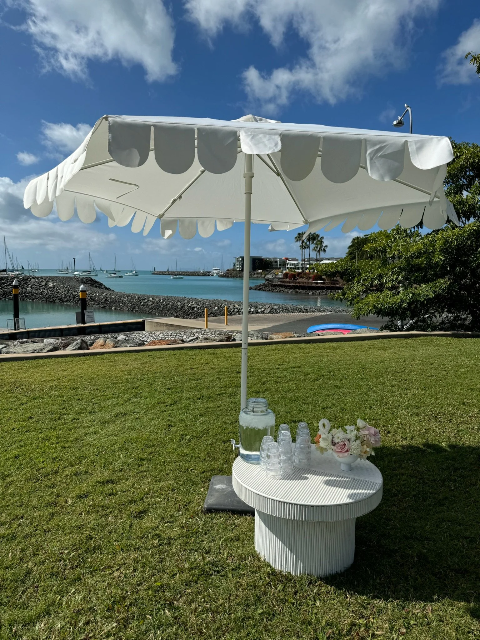 A white outdoor umbrella, a small round white table with stacked cups, a pitcher of water, a flower arrangement, and a glass of water, set on green grass near a body of water with boats, trees, and buildings in the background.