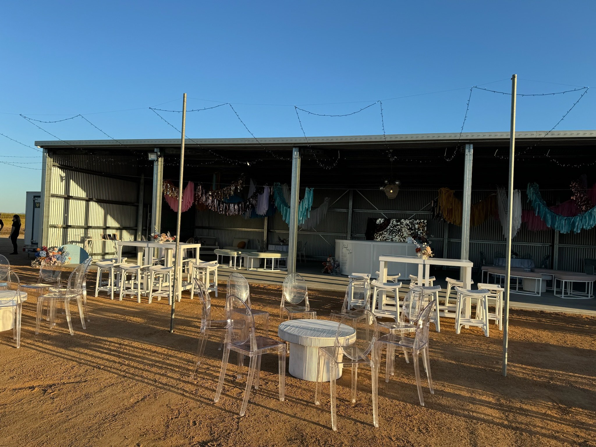 Outdoor event setup with decorated open-air pavilion, transparent chairs, and string lights against a clear blue sky.