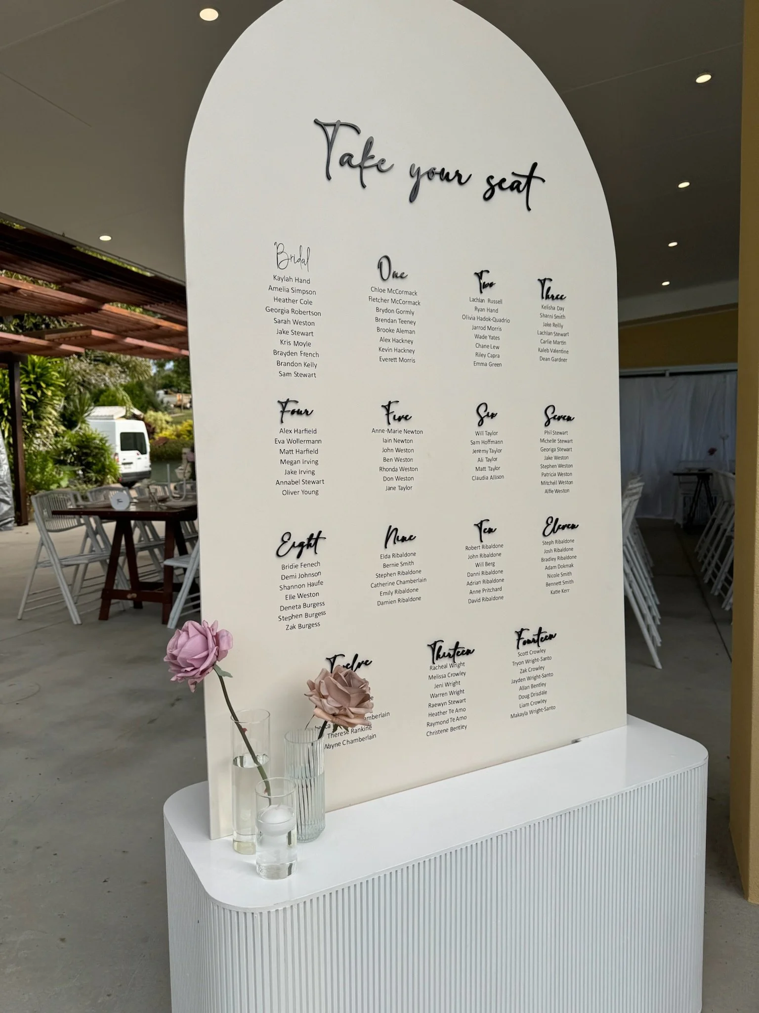 Wedding seating chart titled 'Take your seat' with guest names organized by tables on a white standing board, decorated with pink and purple flowers in vases, placed outdoors with patio furniture in the background.