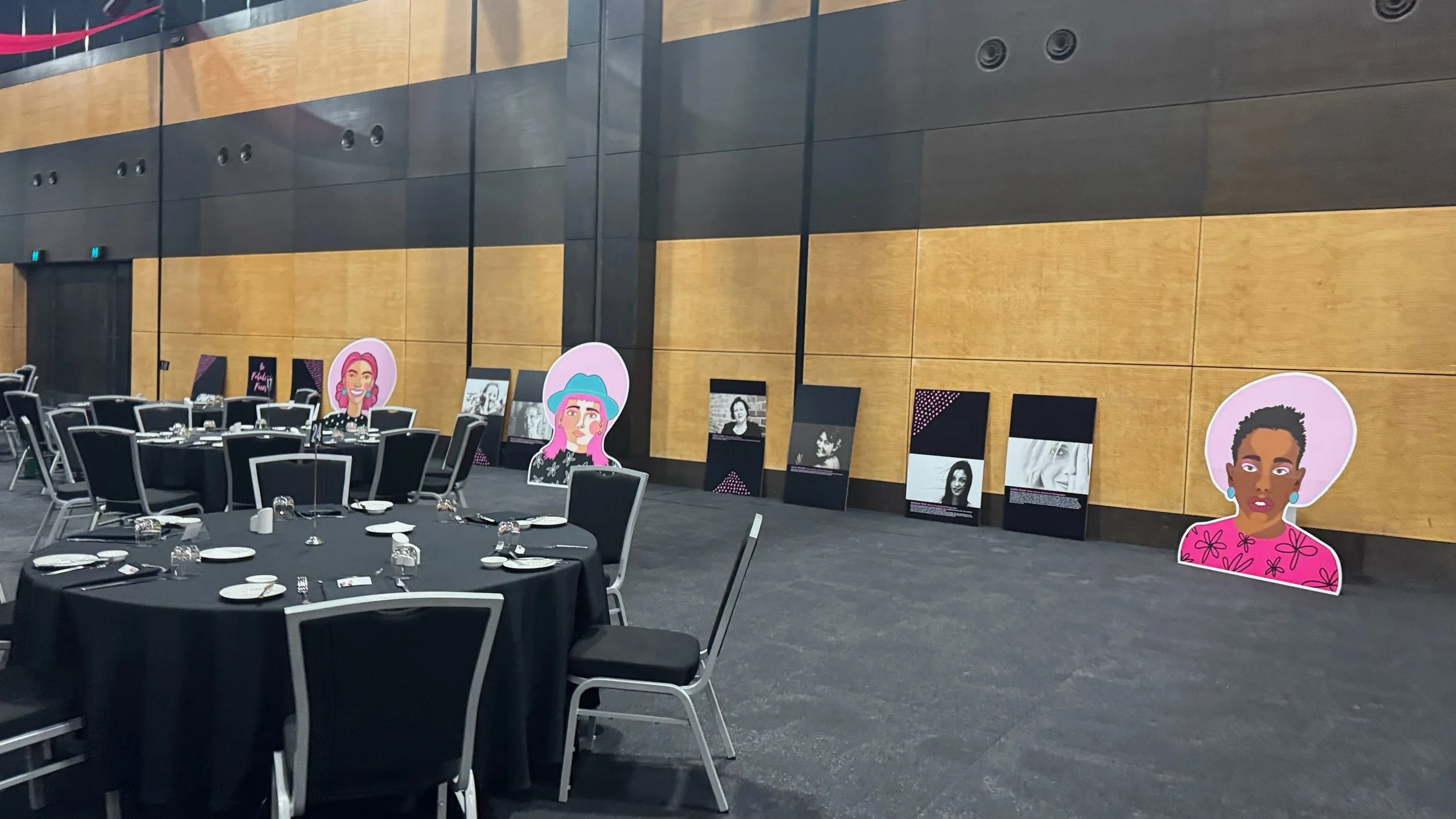 Conference or event hall with black round tables set for a meal, and large colorful illustrations of diverse women displayed along the wall, with some framed photographs of women placed on the floor against the wall.