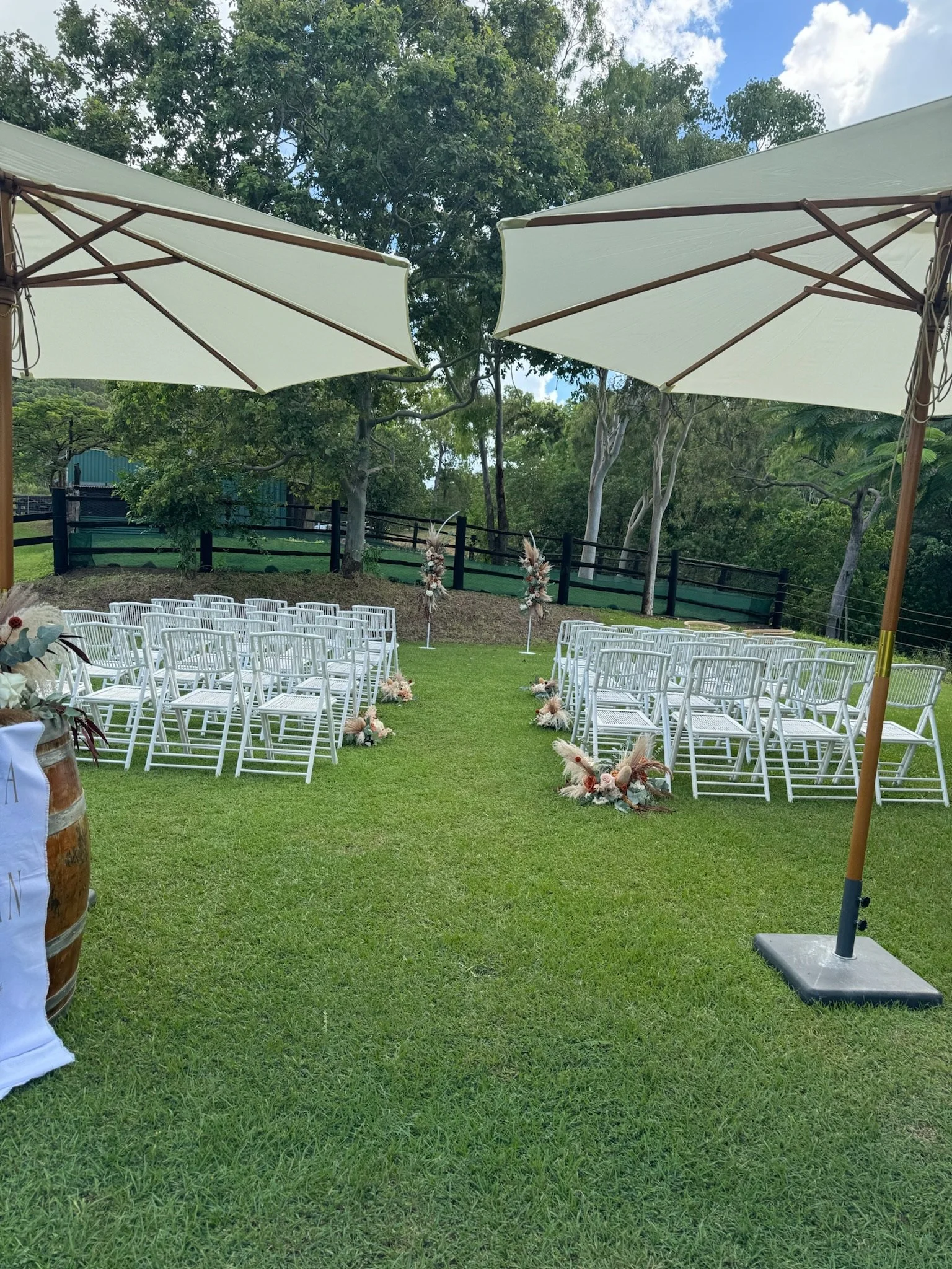 Outdoor wedding setup with white chairs arranged in rows on a grassy area, decorated with floral arrangements, with large white patio umbrellas providing shade, and a backdrop of trees and a fence.