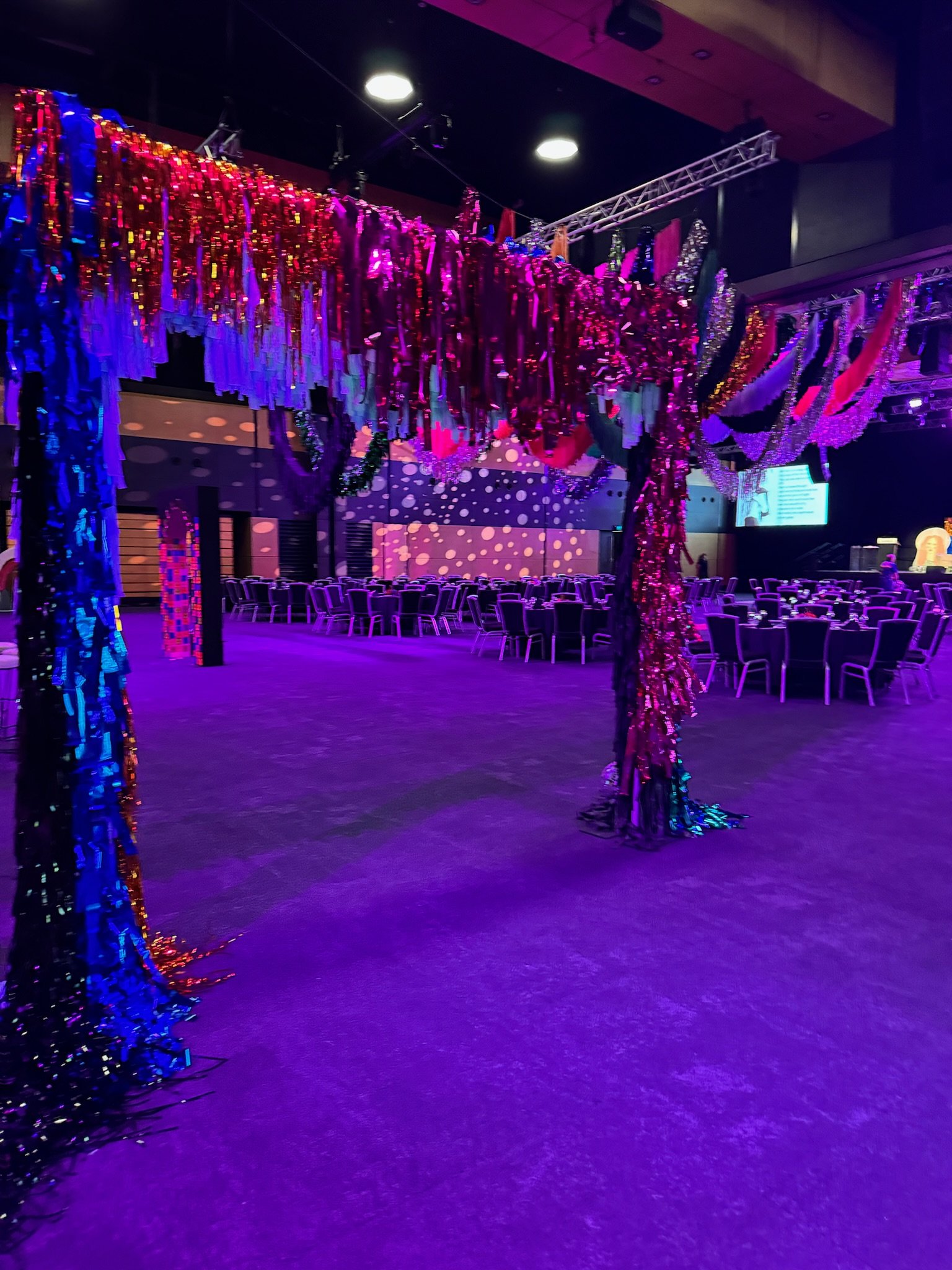 Decorated event space with large, colorful, shimmering hanging installation resembling a tree or canopy, surrounded by rows of chairs and tables, lit with purple and pink lighting.