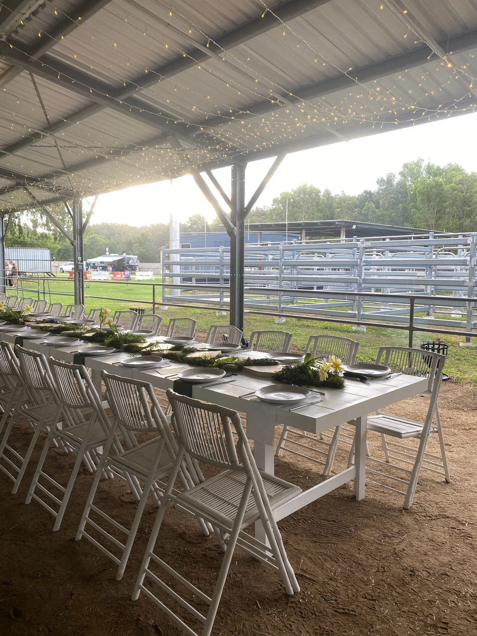 A long white table set with plates, silverware, and floral centerpieces under a canopy with string lights at an outdoor event at a barn or ranch.