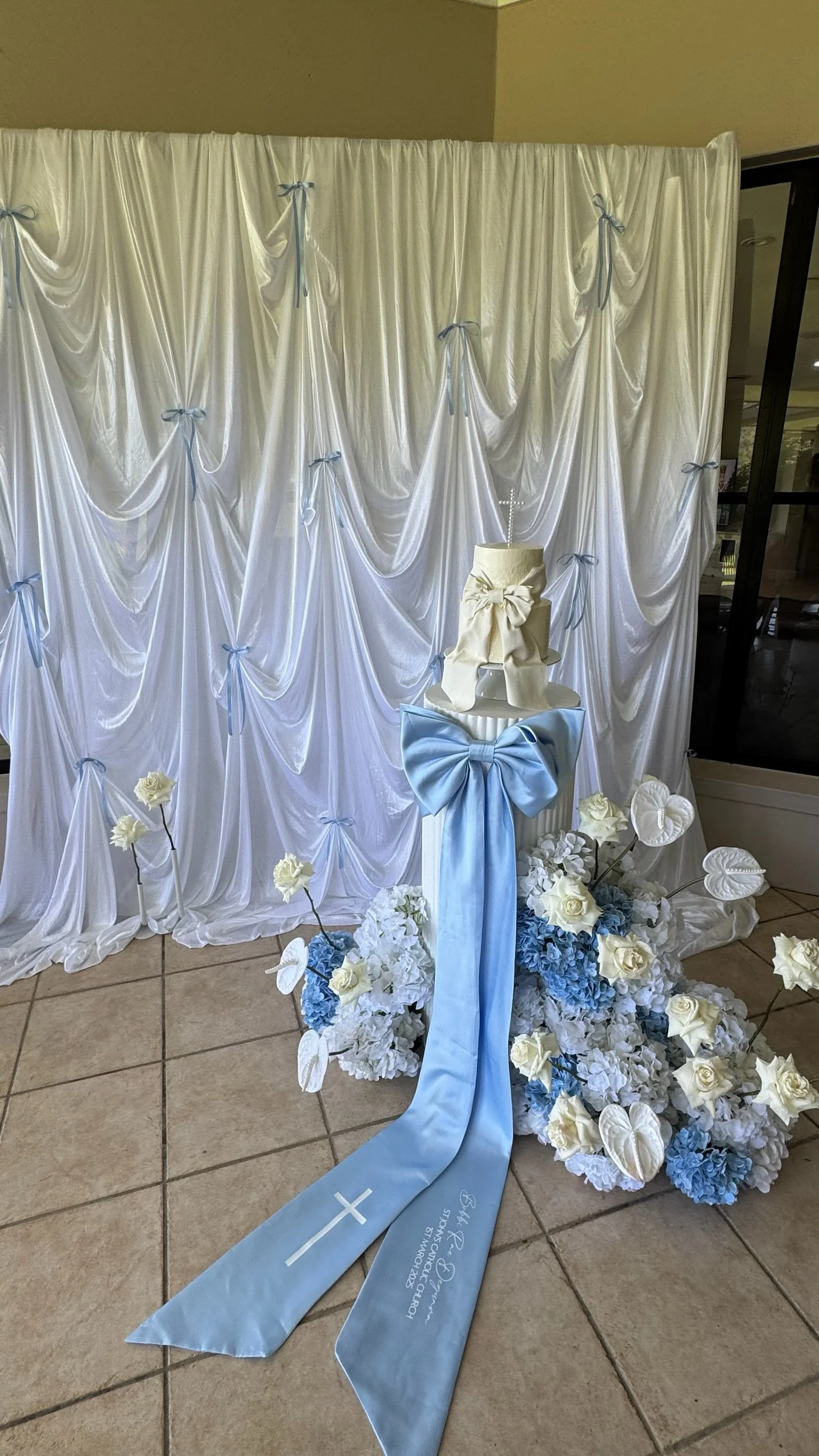 A white and blue floral arrangement with a cake, a large bow, and a ribbon with a cross and name, set against a white draped backdrop tied with blue ribbons, likely for a religious ceremony such as a baptism or first communion.