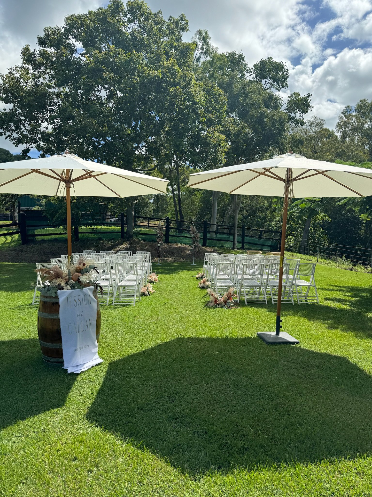 Outdoor wedding or event setup with white chairs arranged on grass, decorated with floral arrangements, two large white umbrellas, a wooden barrel with a sign reading 'Jessica and Callan,' and a scenic backdrop of trees and a partly cloudy sky.
