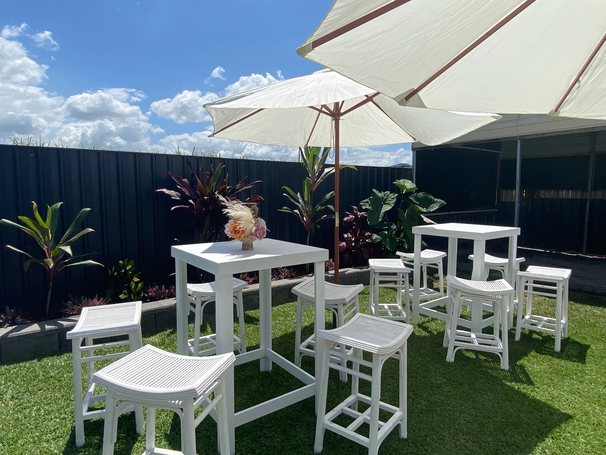 Outdoor patio area with white tables and chairs, large umbrellas, and potted plants against a black fence with a partly cloudy sky.