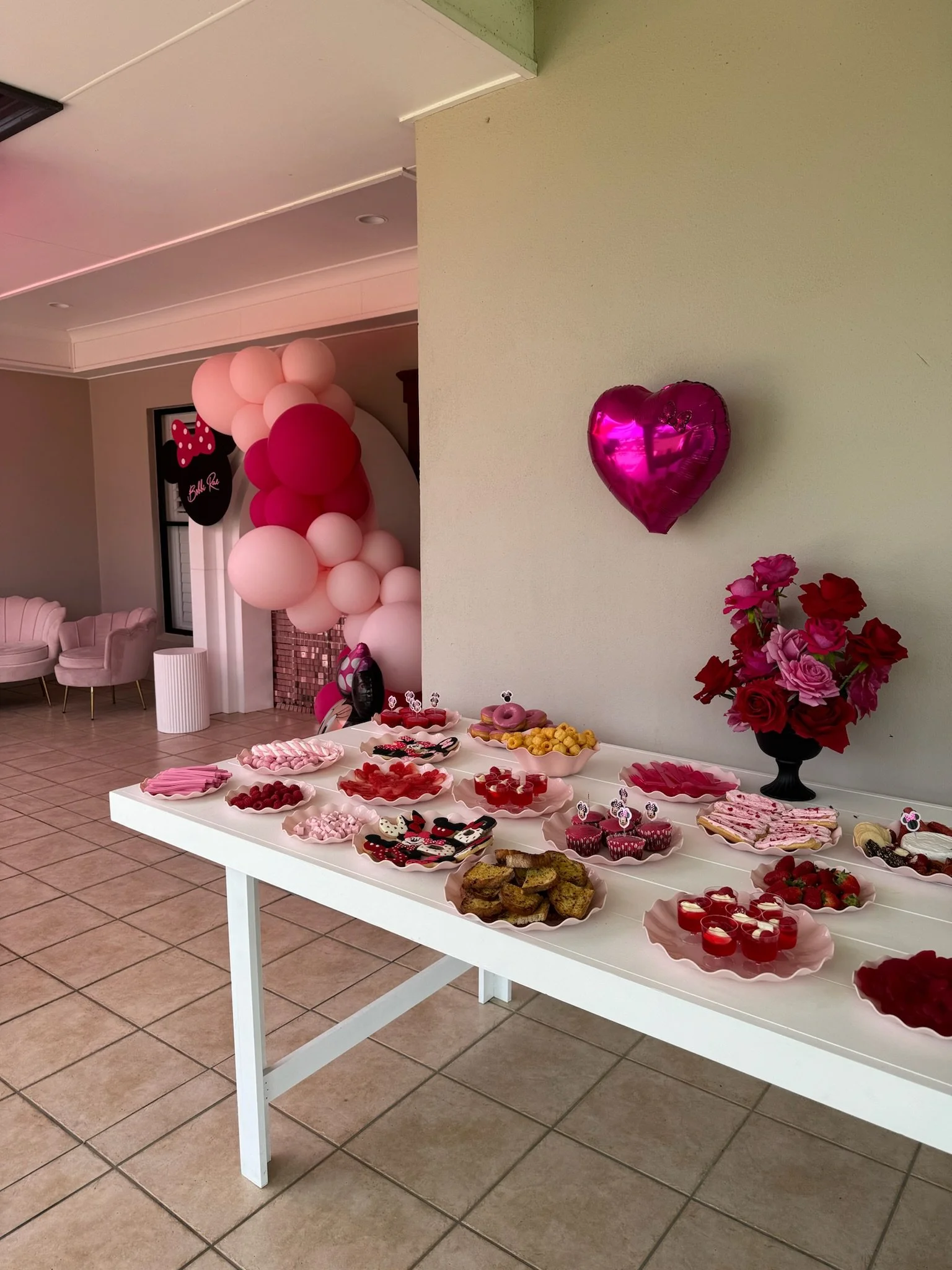 A white table with various pink and red desserts, including cupcakes, cookies, and jello, decorated for a celebration. Pink and red balloons are in the background, along with pink chairs and a floral decoration.