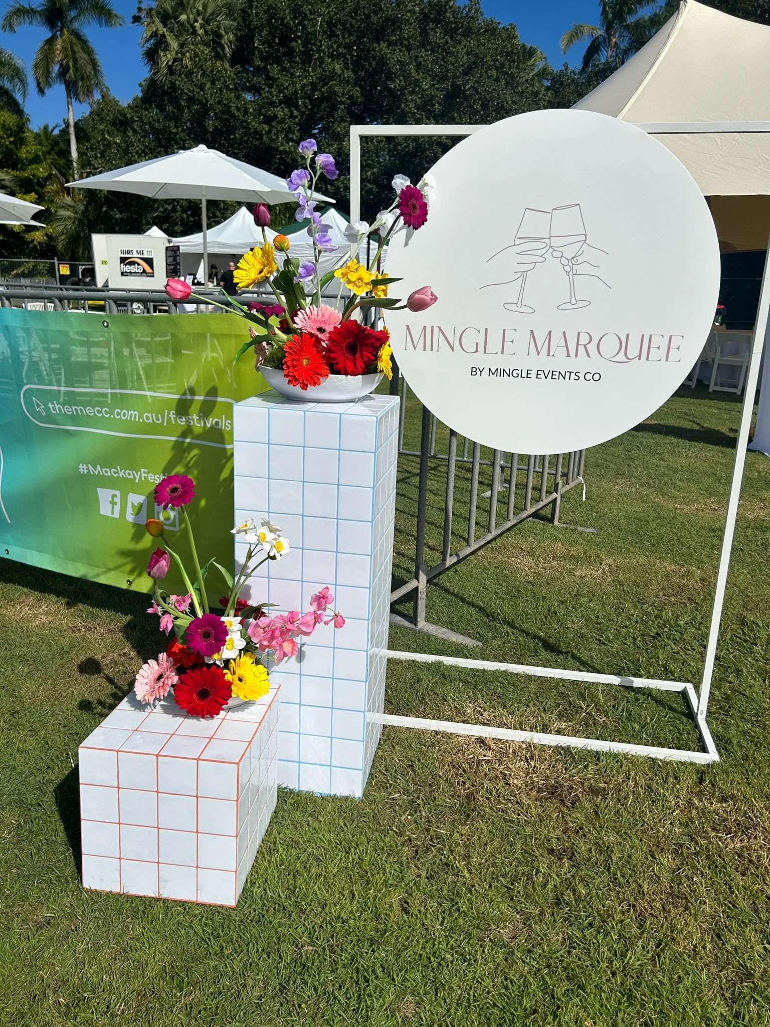 Flowers arranged on white and blue-tiled pedestals in front of a circular sign that reads 'Mingle Marquee by Mingle Events Co' with line art of two people clinking glasses, set outdoors on grass at a festival or event.
