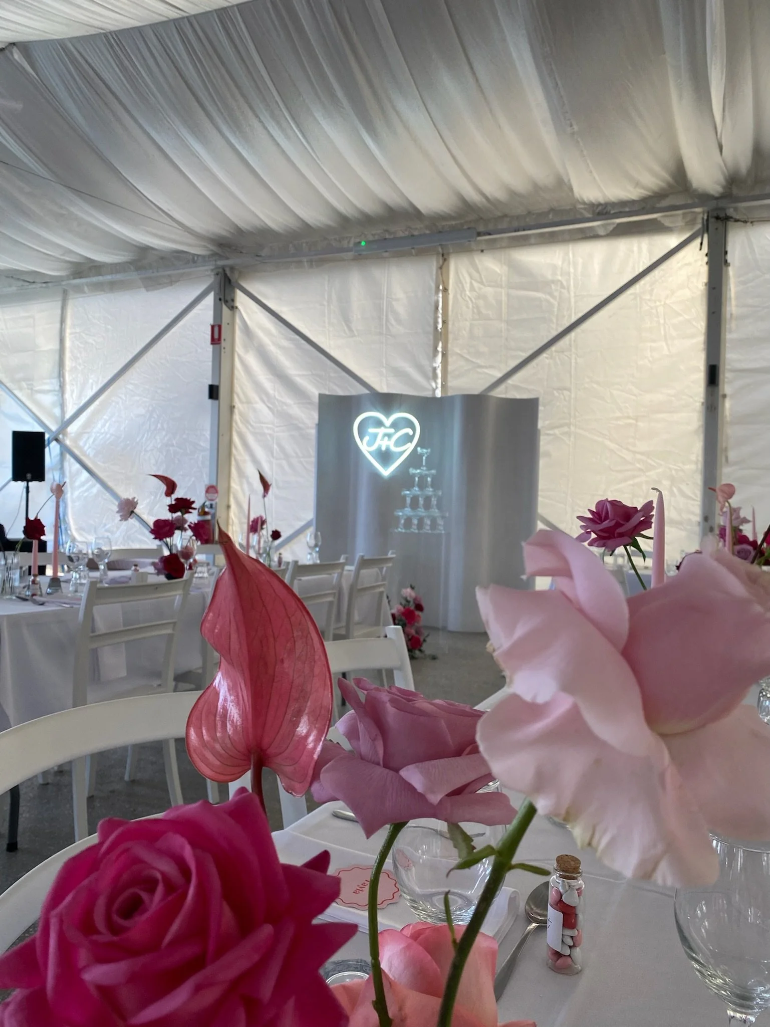 Wedding reception table decorated with pink and red flowers, with a lit-up heart-shaped sign and the letters 'FHC' in the background.
