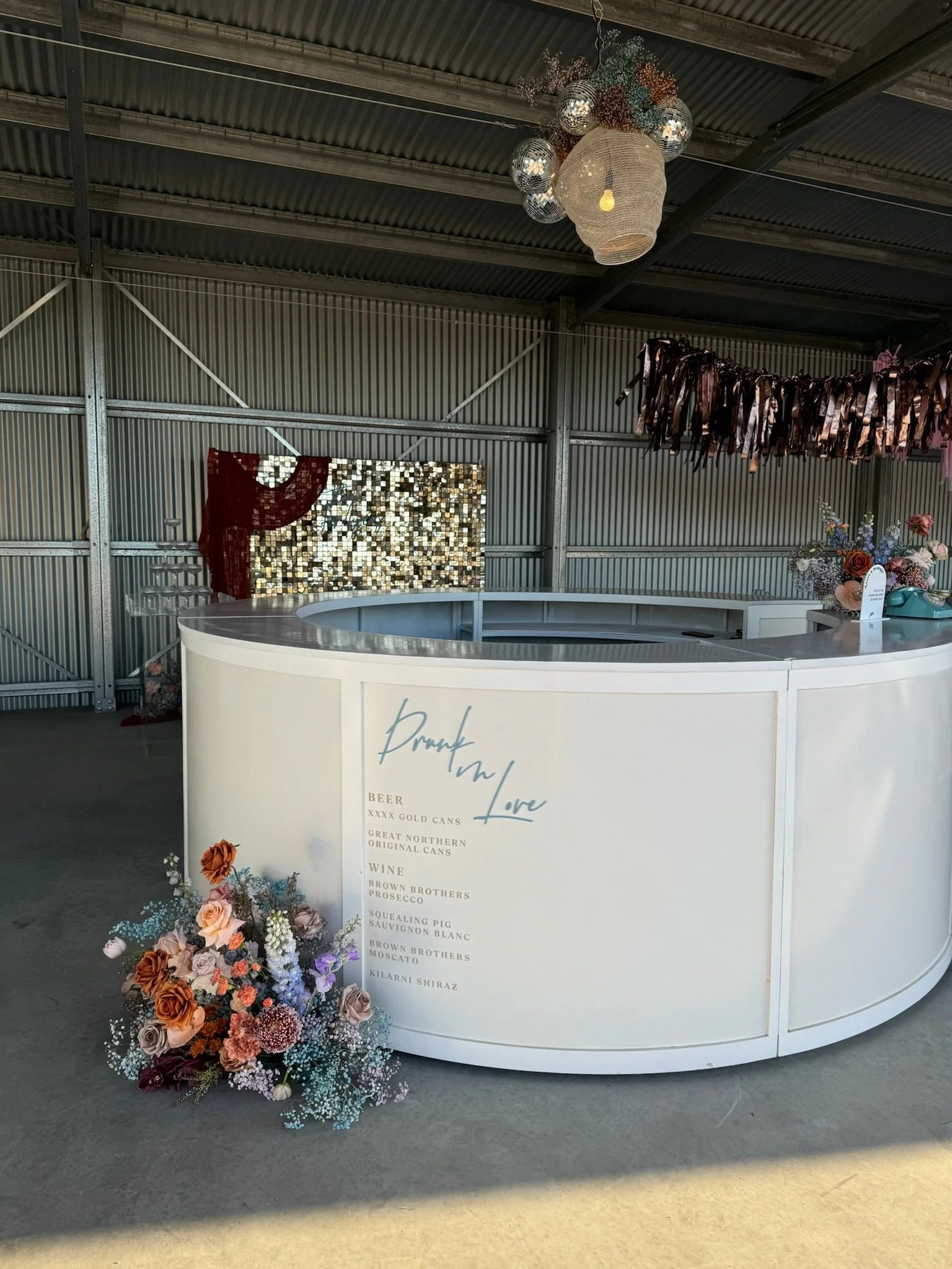 A bar with a white curved counter decorated with a floral arrangement, located in a rustic industrial space with a metal ceiling. Decorations include a large woven face-shaped light fixture, a reflective mosaic backdrop, and hanging metallic and ribb