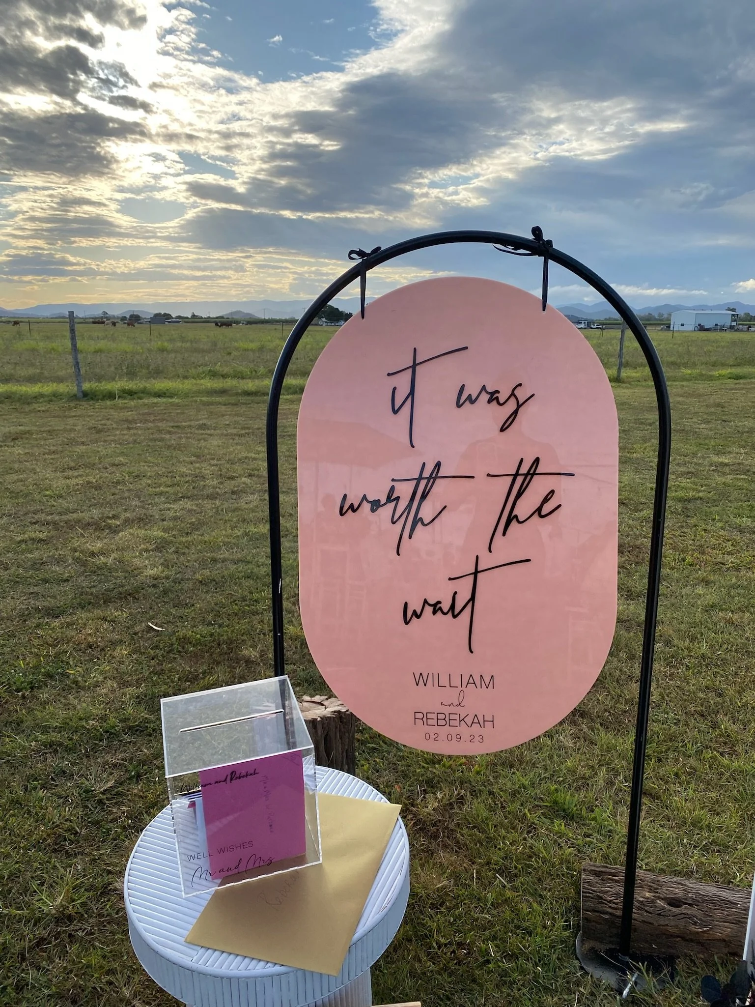 Outdoor wedding sign with the phrase "it was worth the wait" on a pink oval background, with names William and Rebekah and the date 02.09.23 at the bottom. There is a small table with a card and envelopes next to the sign, set on a grassy field with 