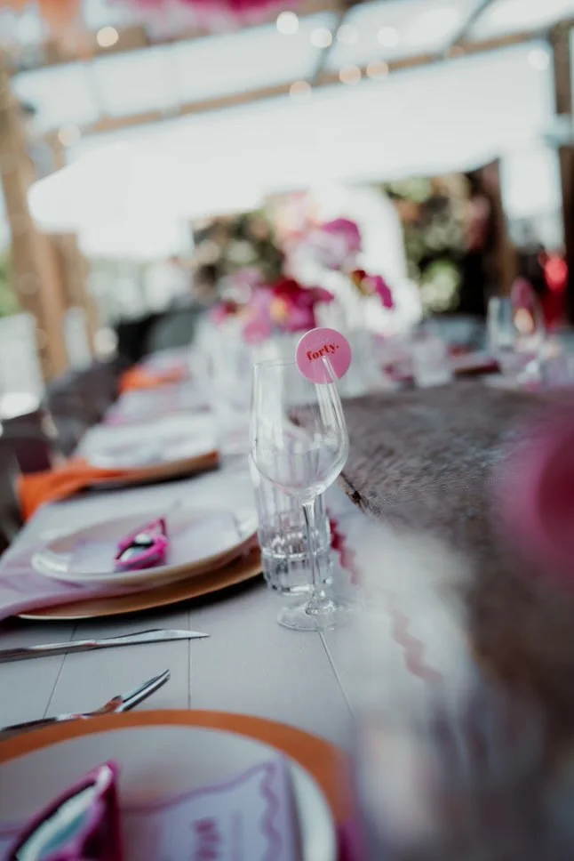 A wedding or event table setting with a glass in the foreground marked with a pink 'Forty!' tag, surrounded by plates, napkins, and utensils; decorated with pink flowers and soft lighting.