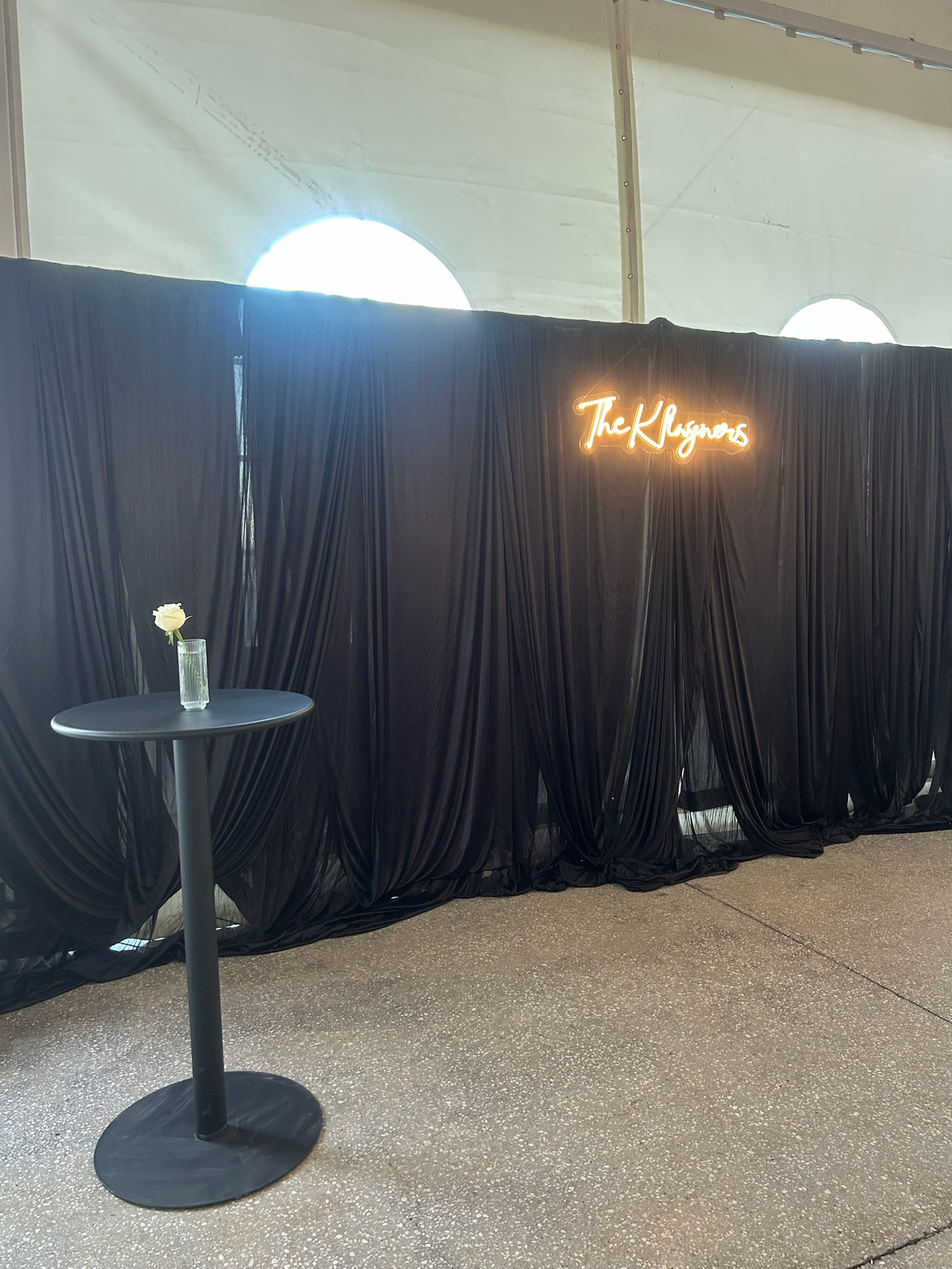 Black curtained backdrop with illuminated neon sign that reads 'The Rhymeers' and a small black round table with a single white flower in a clear glass vase.