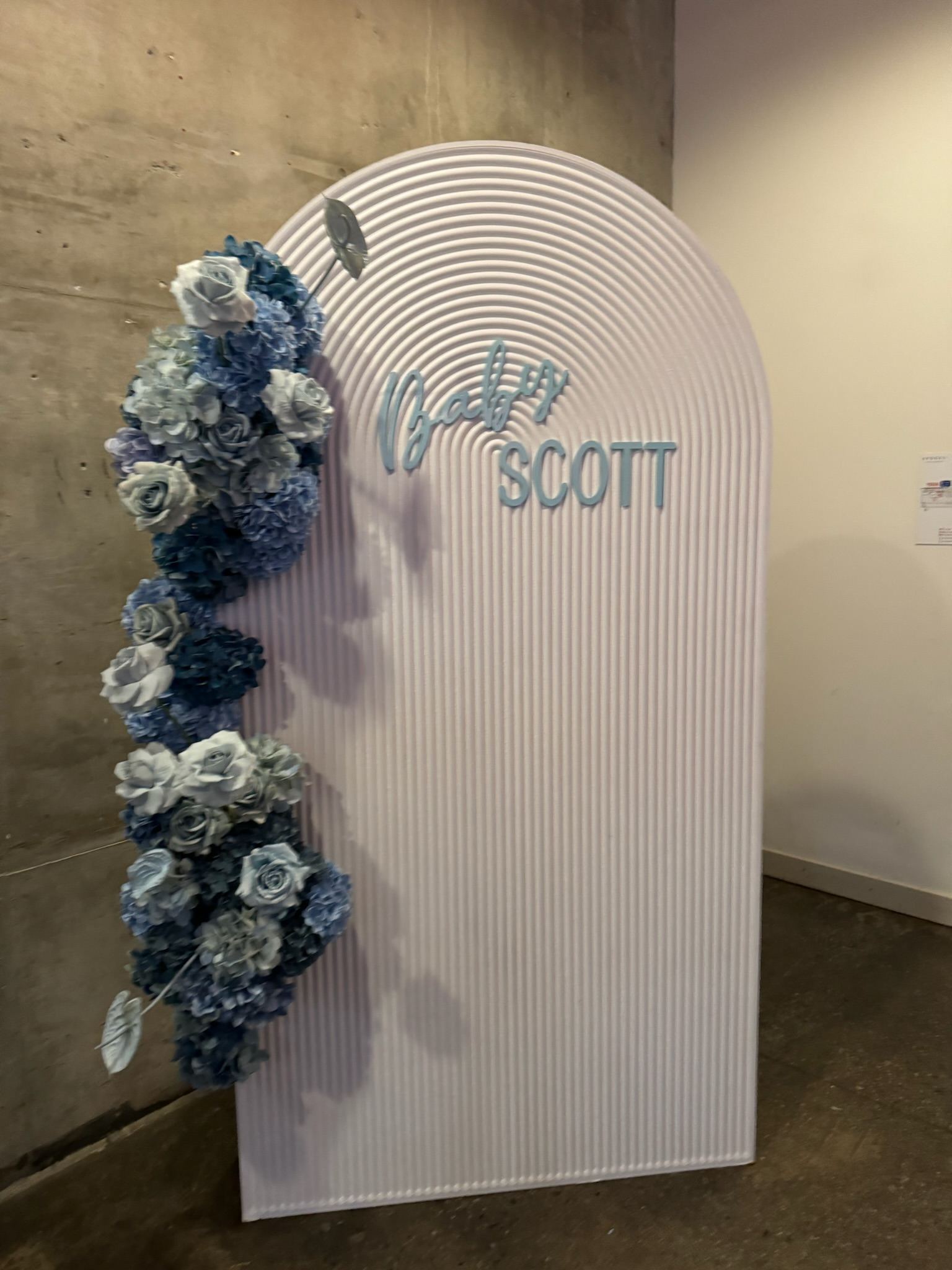 A decorative wedding sign with a white ribbed arch and blue floral arrangement on the left side, displaying the words 'baby SCOTT' in blue cursive and block letters.