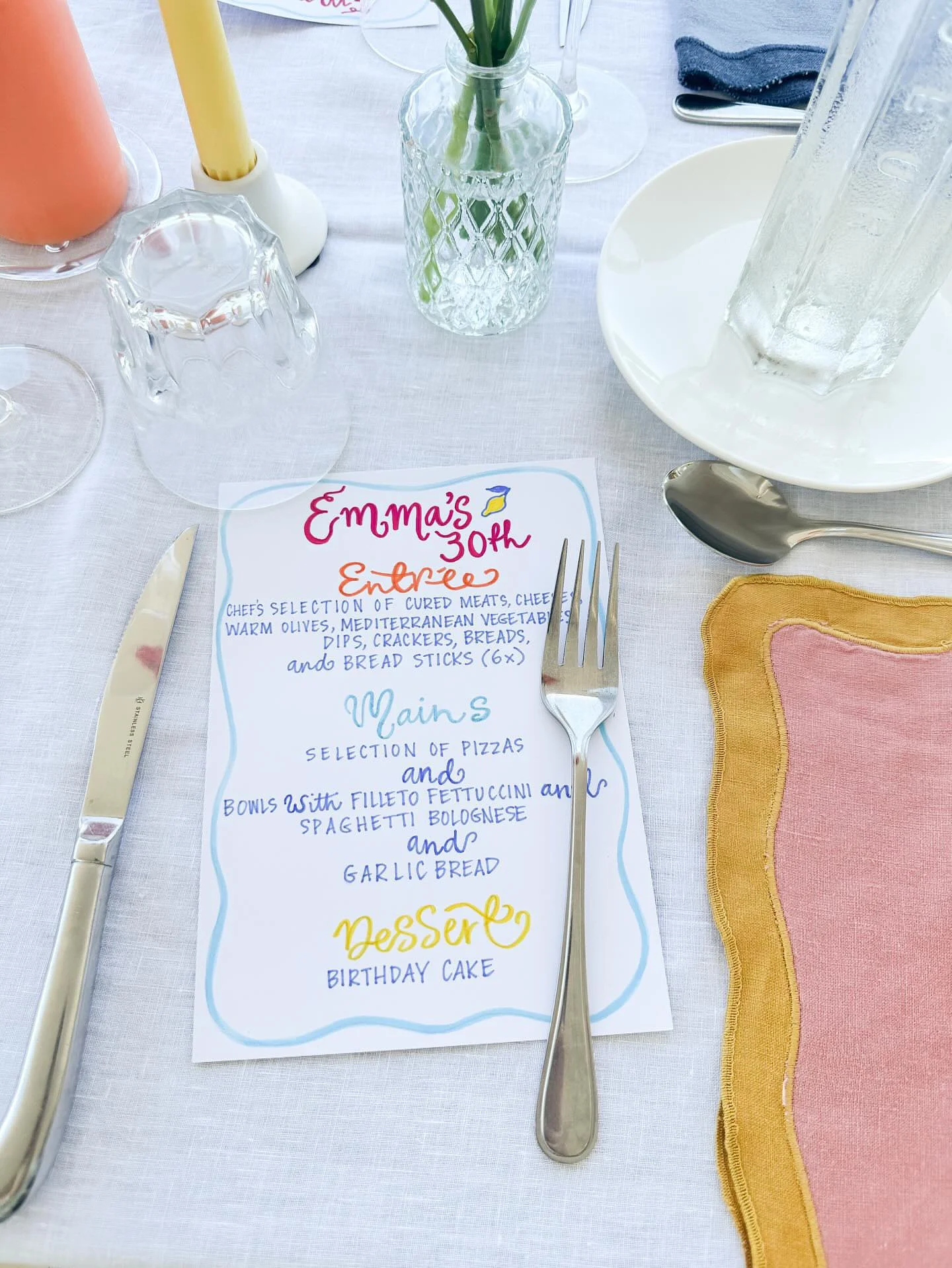A birthday party table setting with a handwritten menu card featuring pink, orange, blue, and yellow text, a fork, a knife, and various glassware, including a vase with flowers and a large glass water pitcher.
