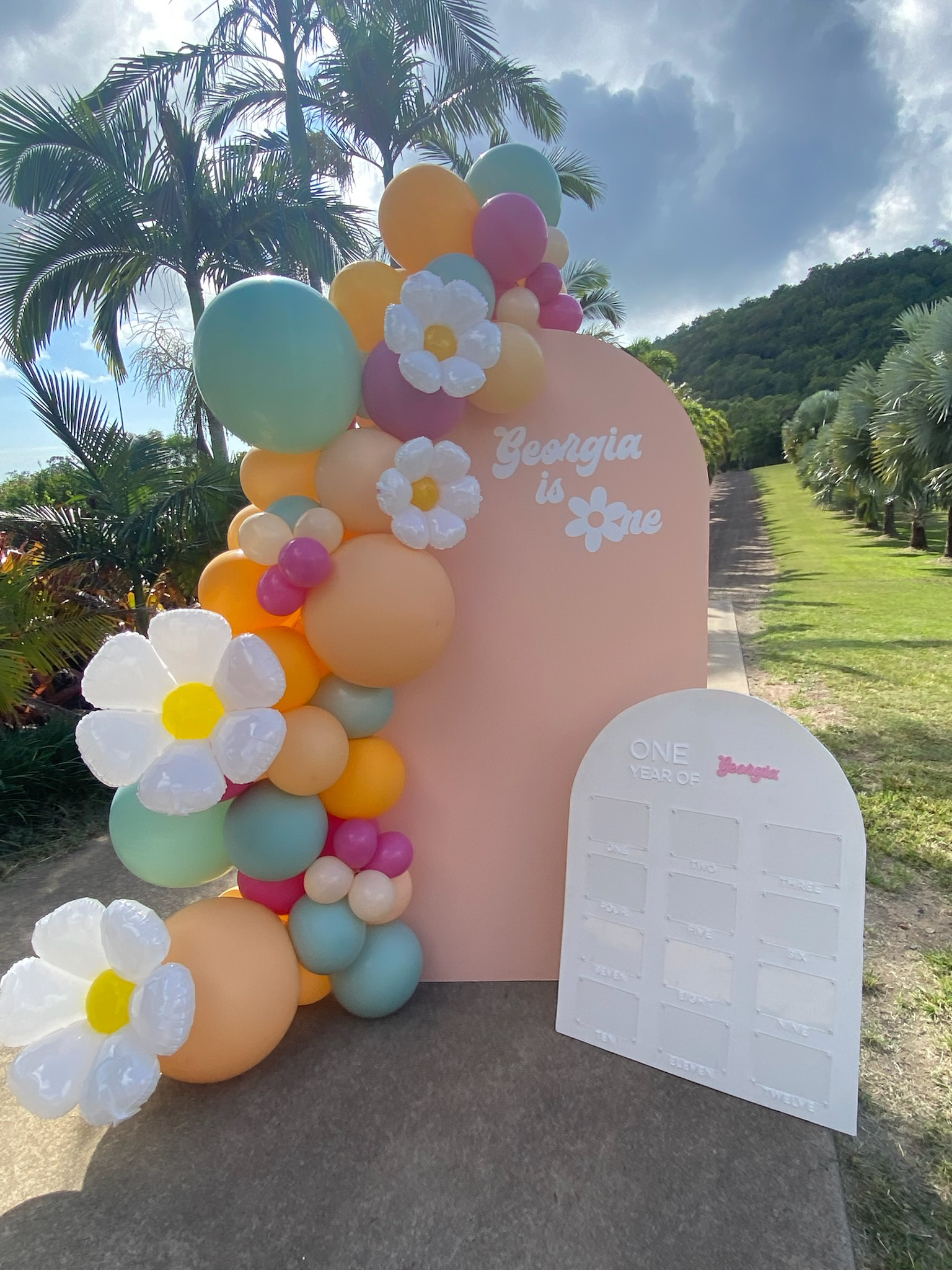 A colorful outdoor display celebrating Georgia's first year. It includes a pink backdrop with balloons in pastel colors, white flowers, and a white board titled "One Year of" with space for writing months. The setting features palm trees and a cloudy