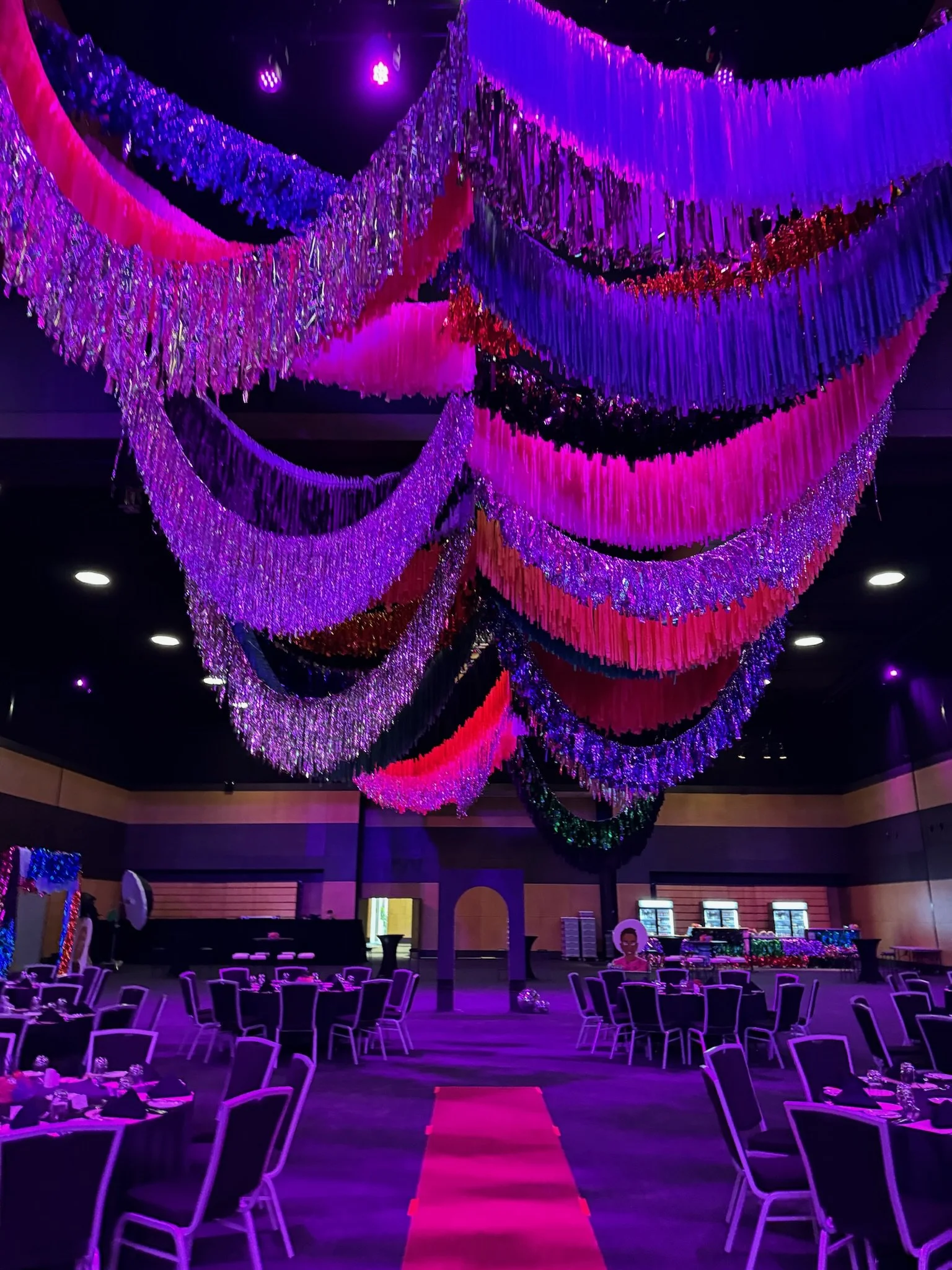 A decorated event hall with colorful hanging drapes and purple lighting, set up for a celebration or party with round tables and chairs, a pink carpet aisle, and a dessert or buffet table in the background.