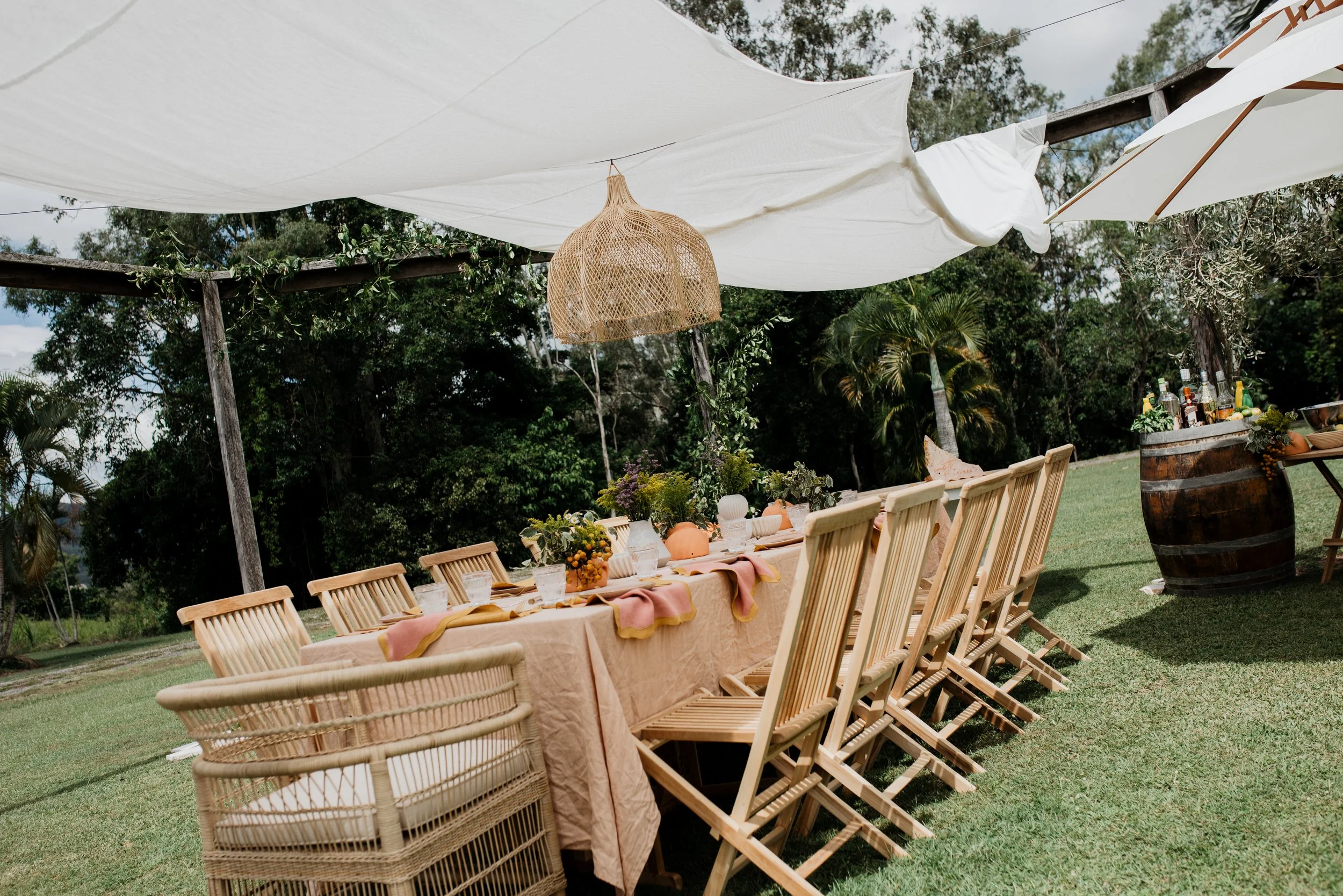 An outdoor dining setup with a long table covered with a beige tablecloth, surrounded by wooden chairs, under a white canopy with a wicker pendant light hanging from it. The table is decorated with flowers and tableware, with a barrel in the backgrou