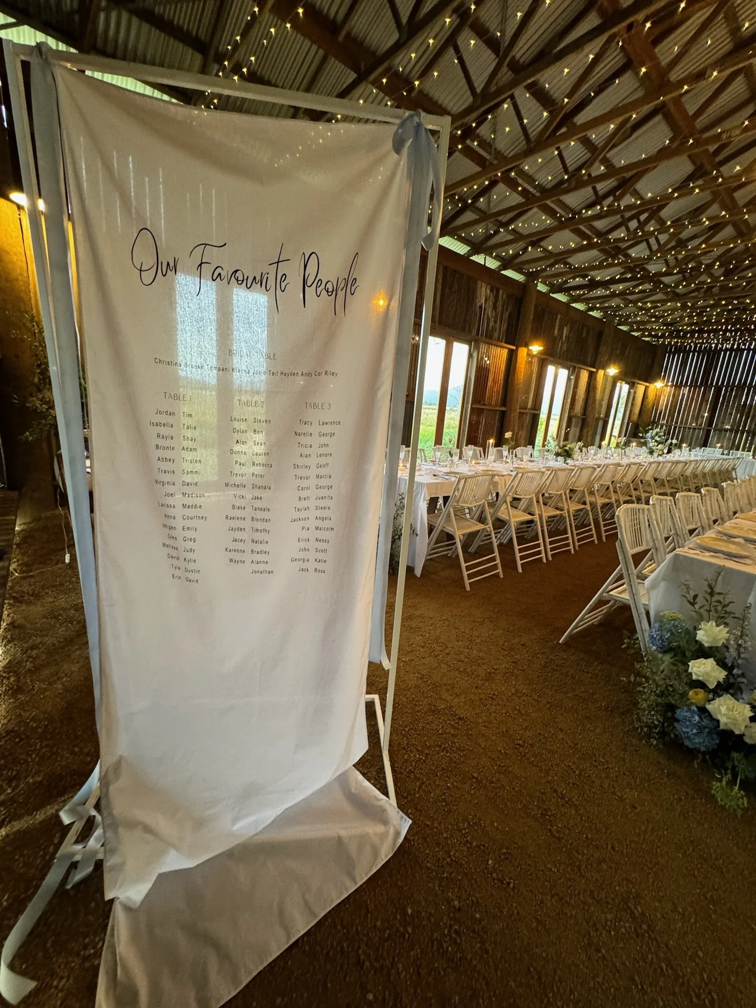 Wedding reception setup inside a barn with string lights, a long table with white chairs, and a large sign listing favorite people.