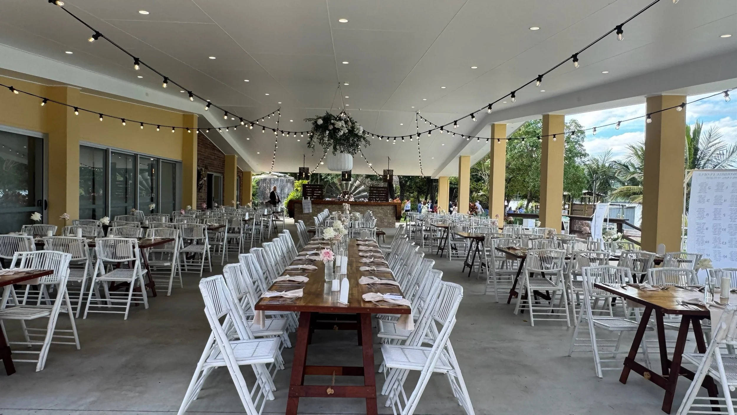 Outdoor event space decorated with hanging string lights, featuring long wooden tables with white chairs and small floral centerpieces, set for a gathering or celebration, with trees and a partly cloudy sky in the background.