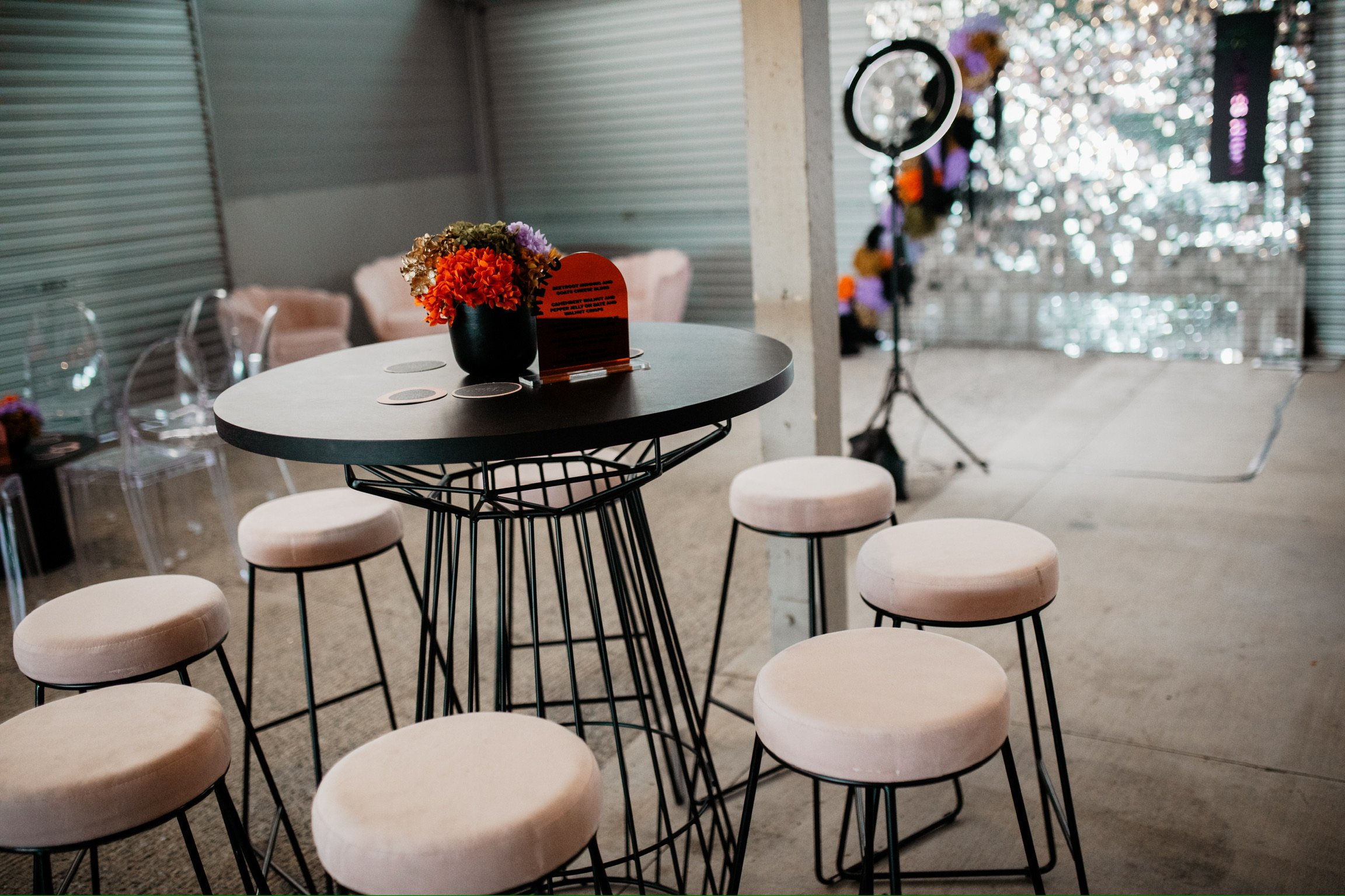A round black table with a bouquet of colorful flowers in a black vase, a small orange sign, and coasters, surrounded by eight beige cushioned stools in a venue decorated with pink, purple, and orange flowers and illuminated by string lights.