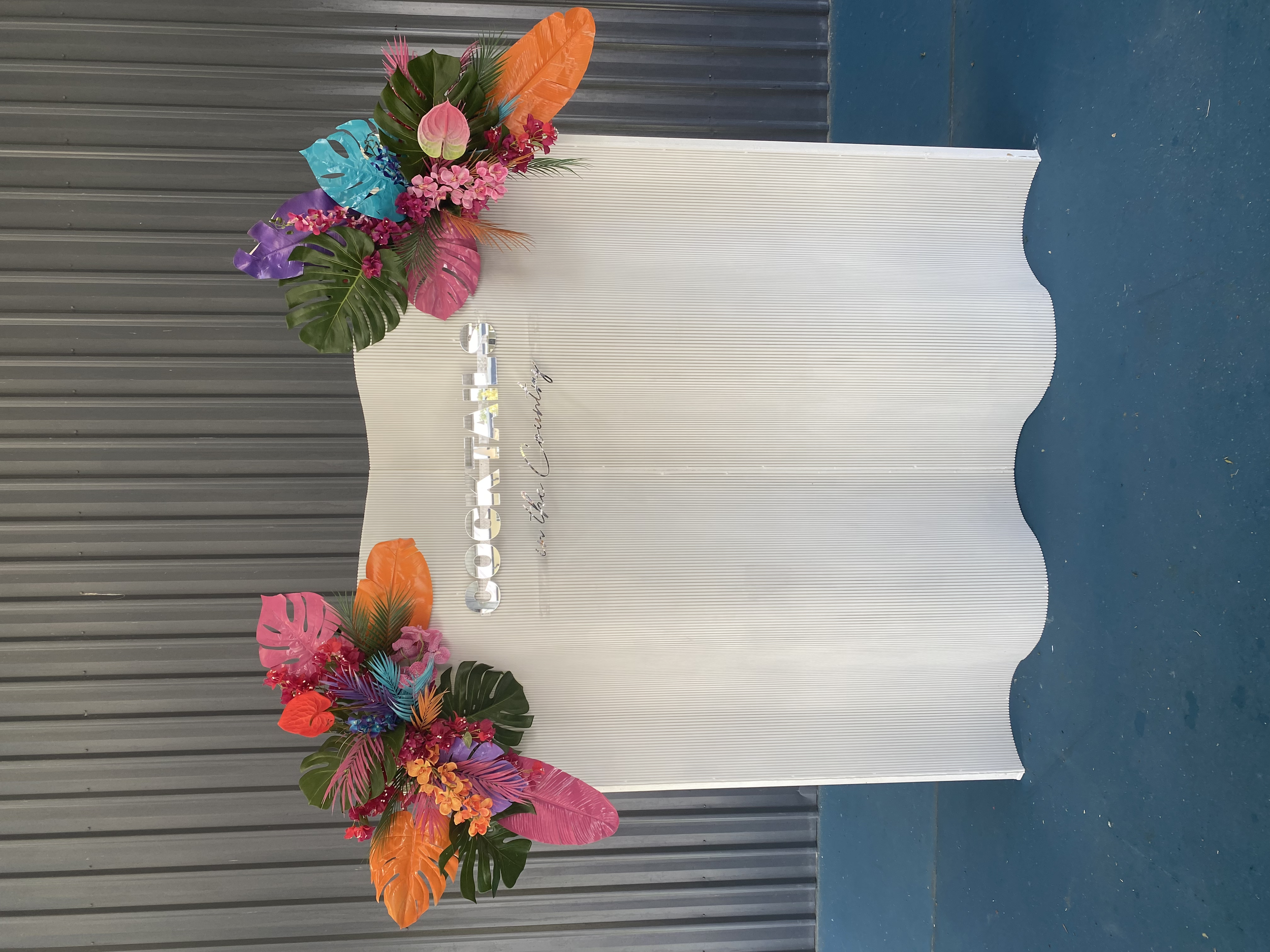 White wavy backdrop with colorful tropical flower arrangements at the top left and bottom right corners, and the word 'Coco' with 'en la Cueva' written underneath in silver and black lettering.