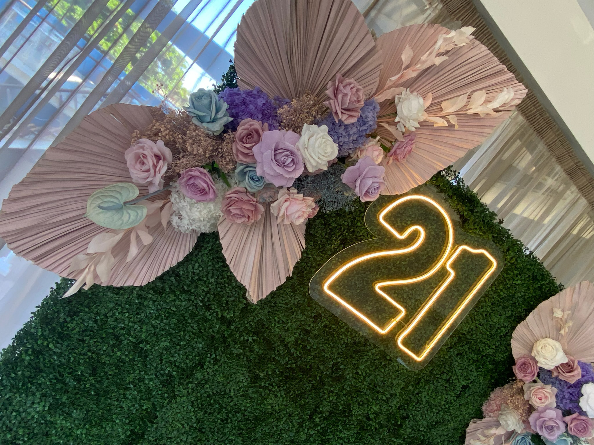 Flower arrangement with pastel roses and hydrangeas on pink paper fans displayed on a green hedge backdrop, with a neon sign showing the numbers 21.