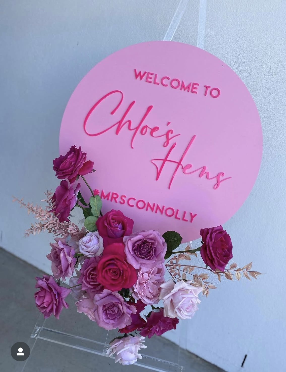A pink sign with the text "Welcome to Chloe's Hens" and the hashtag "#MRSCONNOLLY," decorated with a bouquet of pink, purple, and white roses.