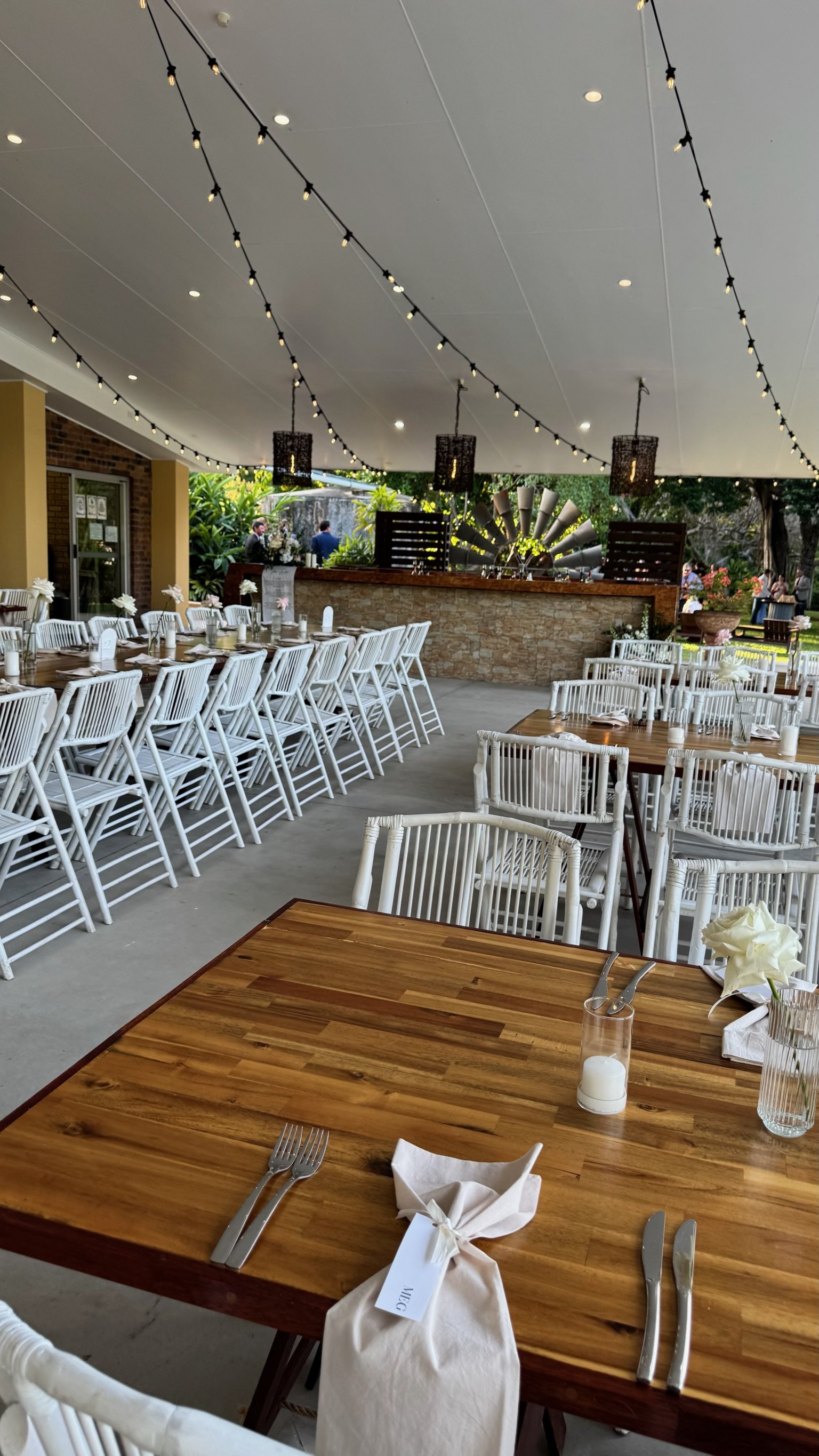 Decorated outdoor restaurant dining area with wooden tables, white chairs, string lights on the ceiling, and a stone bar with a windmill decoration in the background.