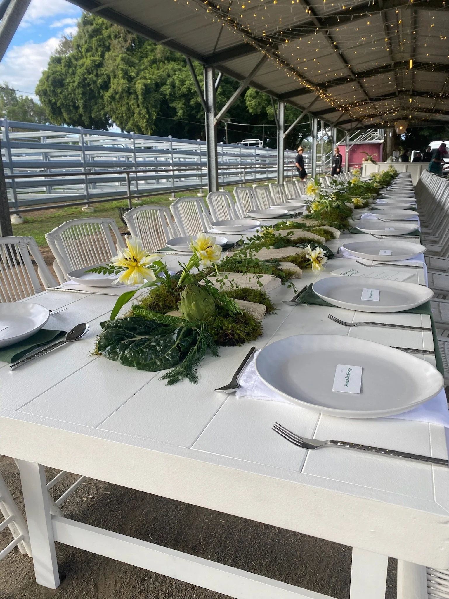 Long outdoor banquet table set with white plates, silverware, and floral centerpieces under a covered area decorated with string lights.
