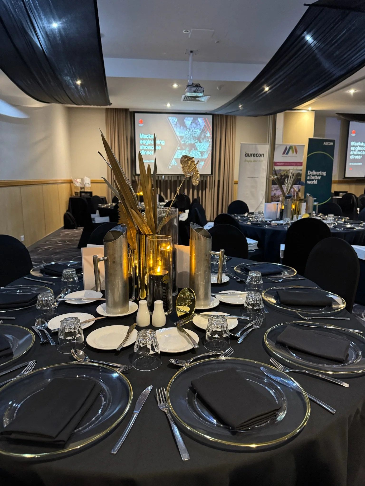 Round banquet table set for a formal event with black tablecloth, black napkins, silverware, glassware, and white plates, with gold and black centerpieces, in a decorated event hall with large screens and banners in the background.