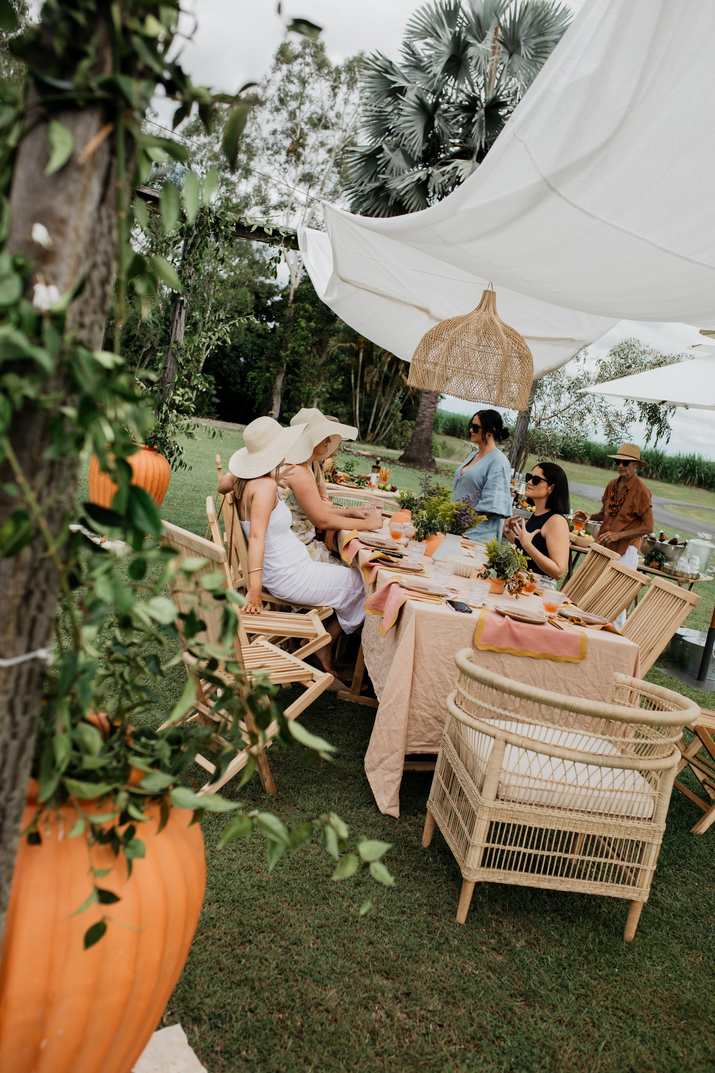 People gathered around an outdoor dining table with peach-colored tablecloths, under white canopy, in a lush garden setting with trees and large potted plants.