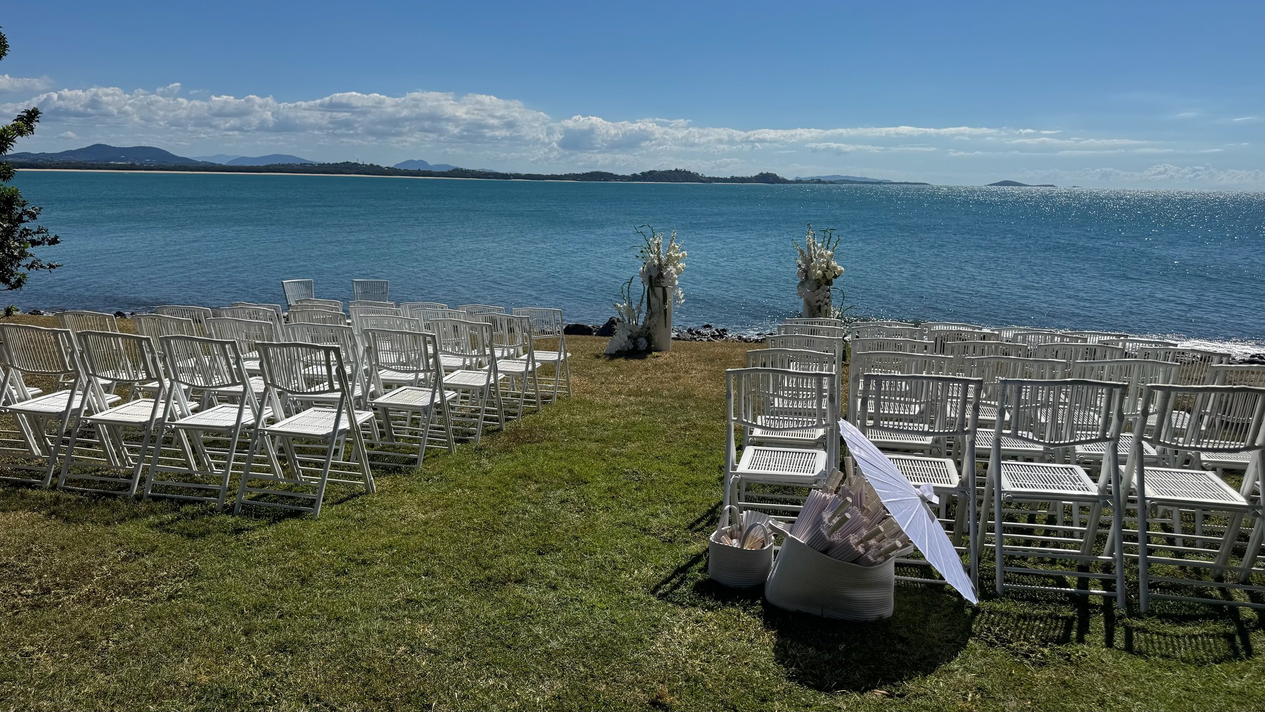 Outdoor wedding setup on a grassy area by a large body of water, with white chairs arranged facing an aisle, floral arrangements, and a decorative umbrella
