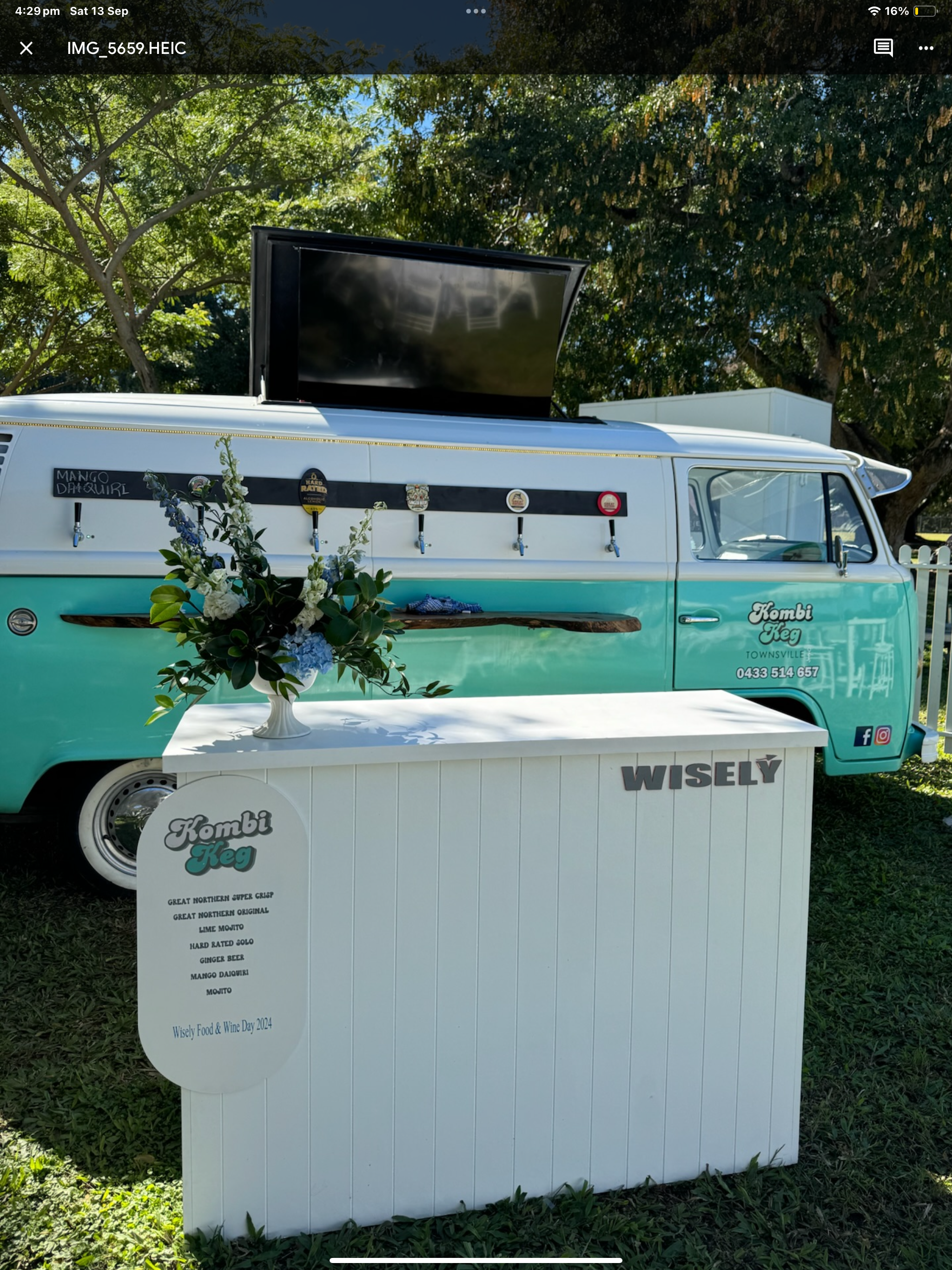 A vintage teal and white camper van with a black stripe, parked outdoors under trees. The van has a flat-screen TV on its roof and several novelty beer taps mounted on the side. An informational sign in front displays the menu for "Kombi Keg" drinks,