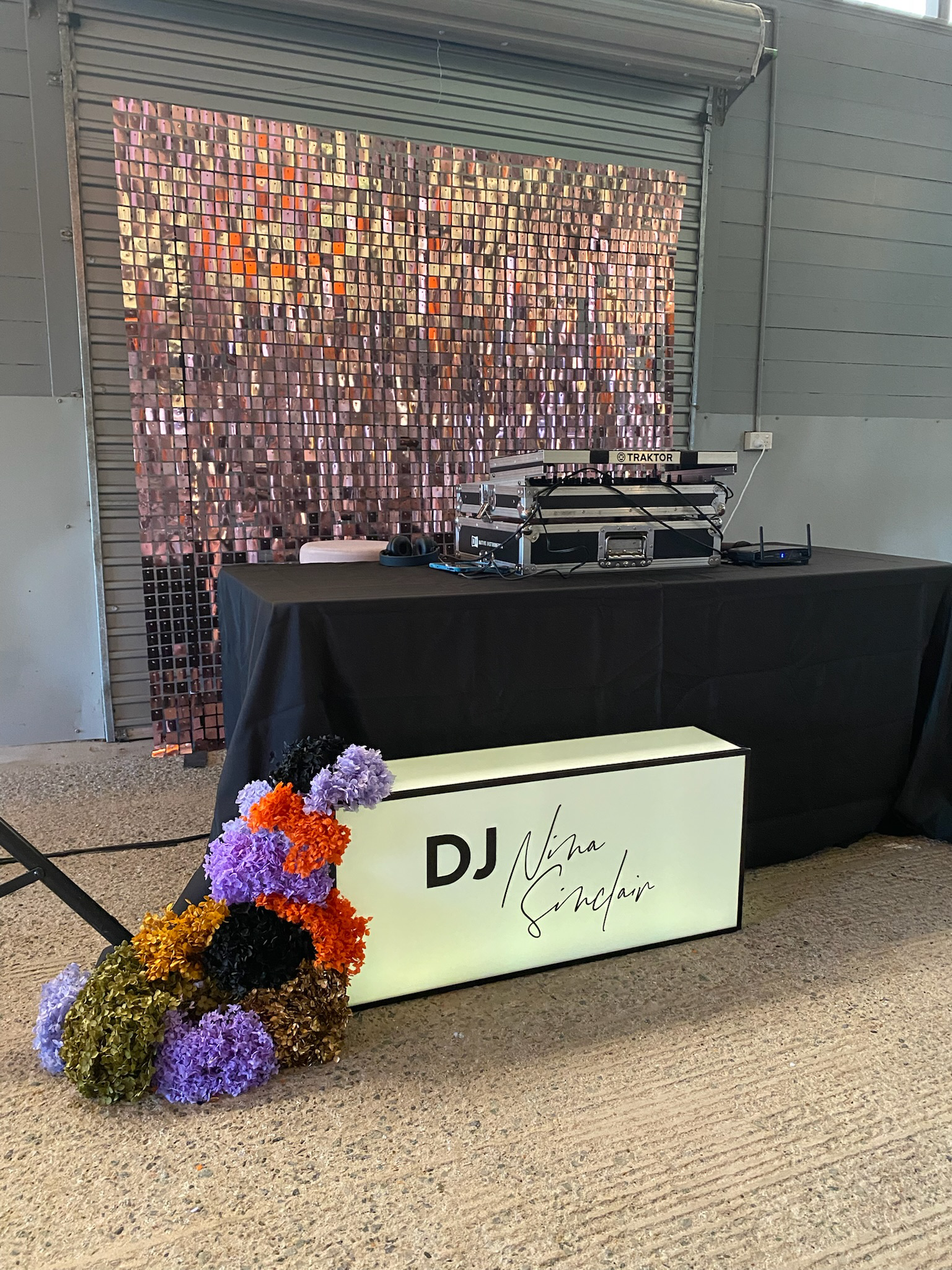 A DJ setup at an event, with a sign that reads 'DJ Niam Sinclair', a floral arrangement of purple, orange, black, and brown flowers, a black tablecloth, and a glittery pink backdrop behind the DJ equipment.